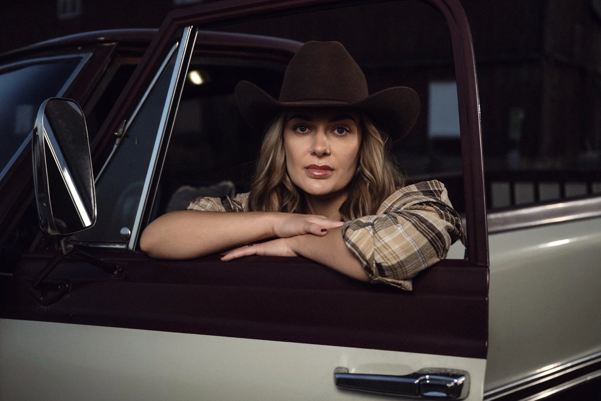Kvinna med en cowboyhatt som sitter i en bil, tittar mot kameran, med armar vilande på fönsterkarmen.