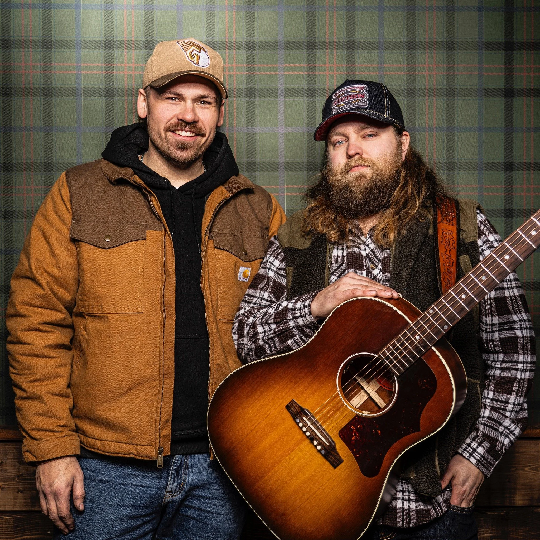 Två män, en med gitarr, står framför ett grönt rutigt tapet.