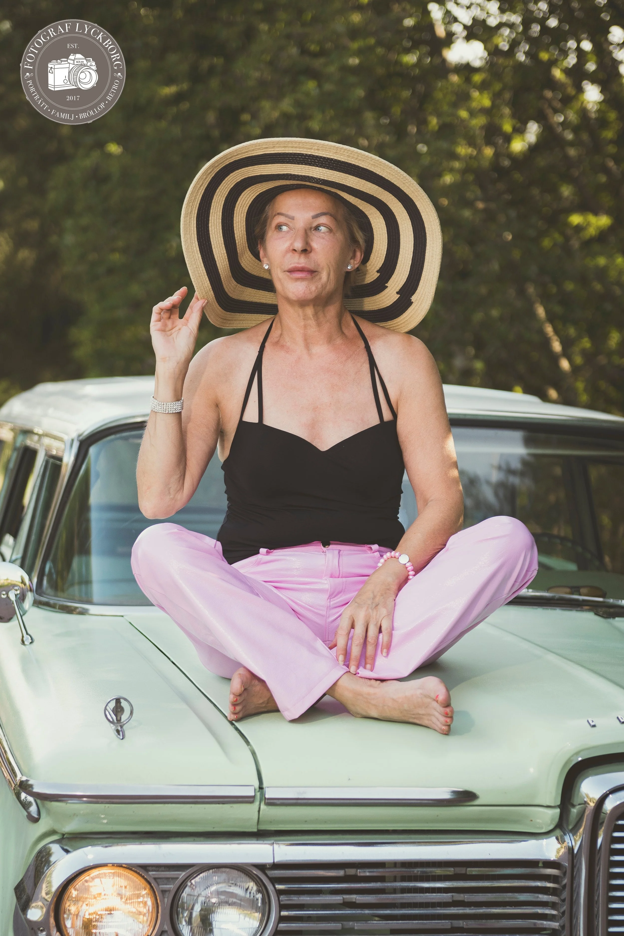 En äldre kvinna i svart topp och rosa byxor sitter med korslagda ben på motorhuven av en ljus grön bil, med en stor bredrutig hatt och håller i kanten av hatten. Bakgrunden är en gröngräset skog.
