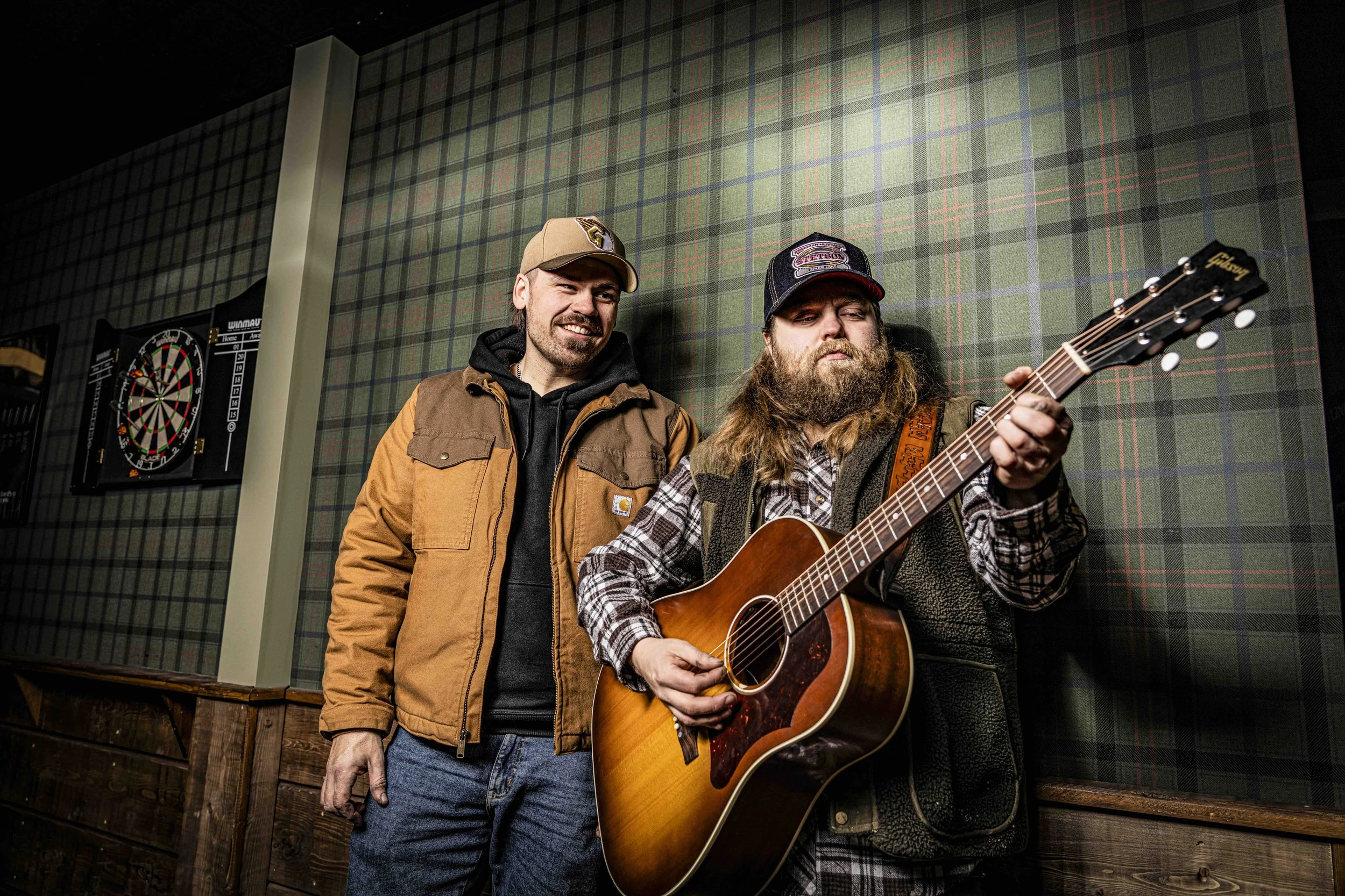 Två män, en spelar gitarr och den andra lyssnar, inomhus mot en vägg med tartanmönster.