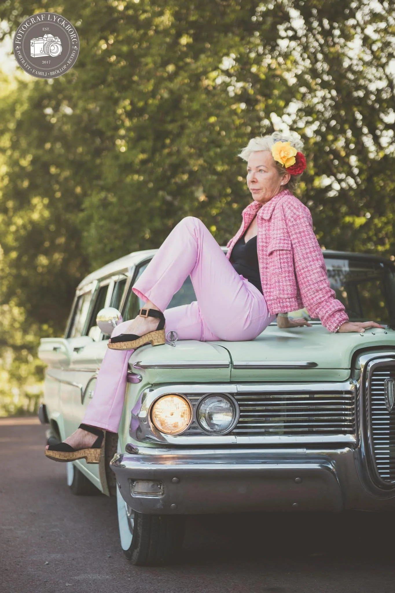 En äldre kvinna sitter på motorhuven till en bil i ett naturskönt område med många gröna träd. Hon bär en rosa kavaj, rosa byxor, svarta sandaler och har en blomsteraccessoar i håret.