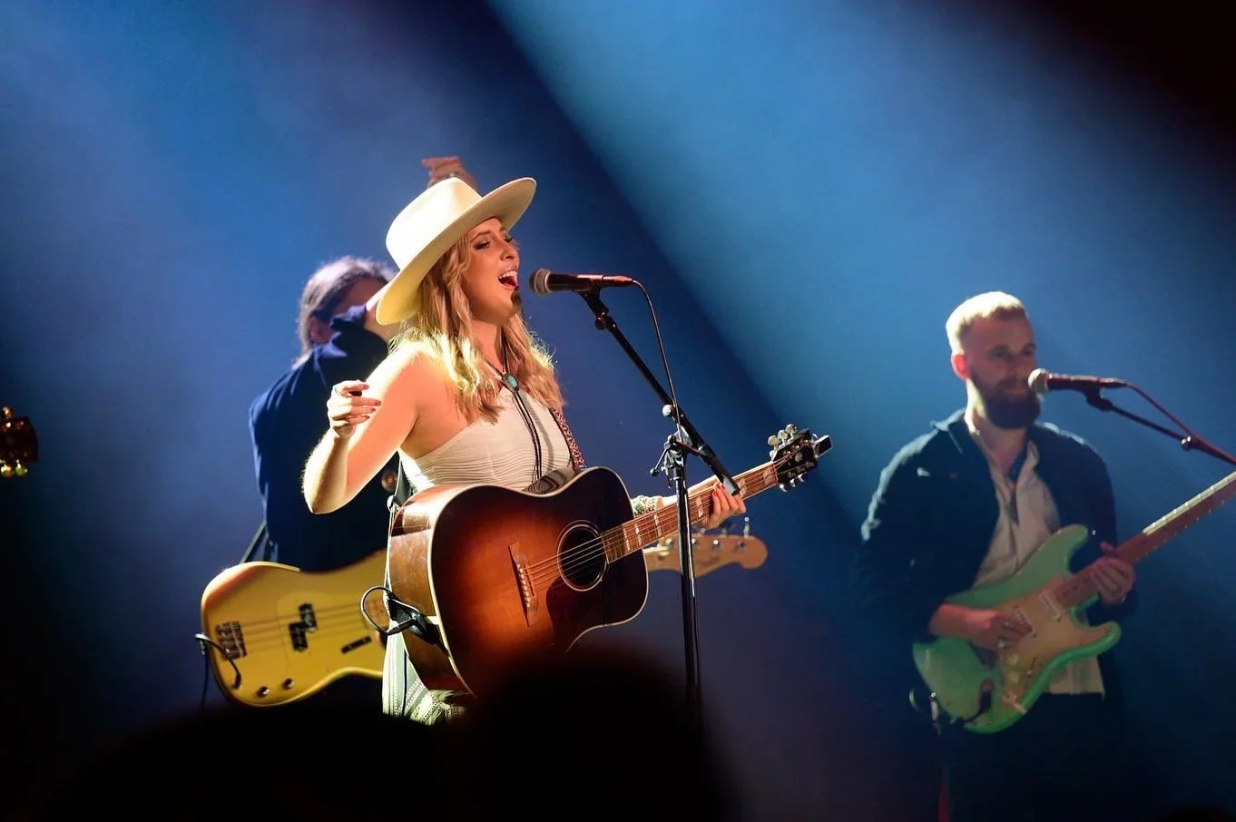 En kvinna som sjunger och spelar akustisk gitarr på scen med två män som spelar elgitarr och bas i bakgrunden.