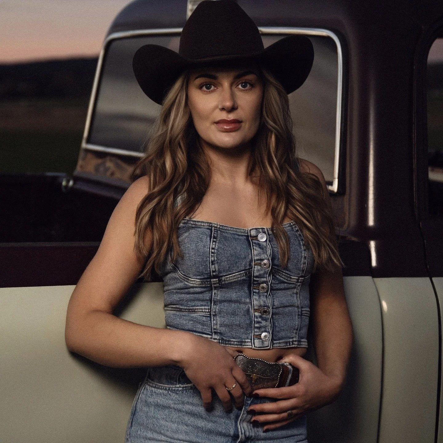 Kvinna i cowboyhatt och jeansoutfit står framför en bil, kvällstid.