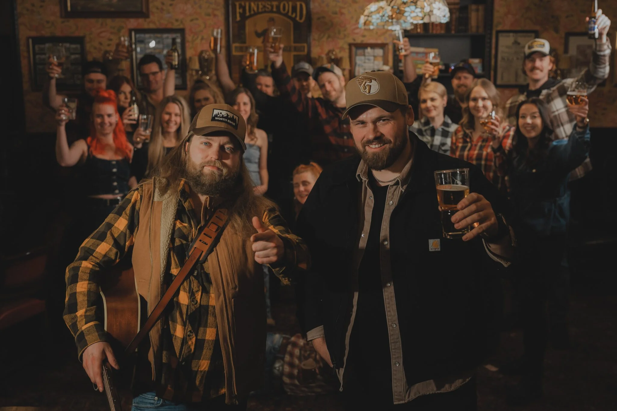 En grupp människor som firar på en bar, många håller glas med öl och vinkar. Två män i förgrunden, en med gitarr och en annan med ett ölglas, båda skrattar. Bakgrunden visar en vägg med bilder och tavlor.