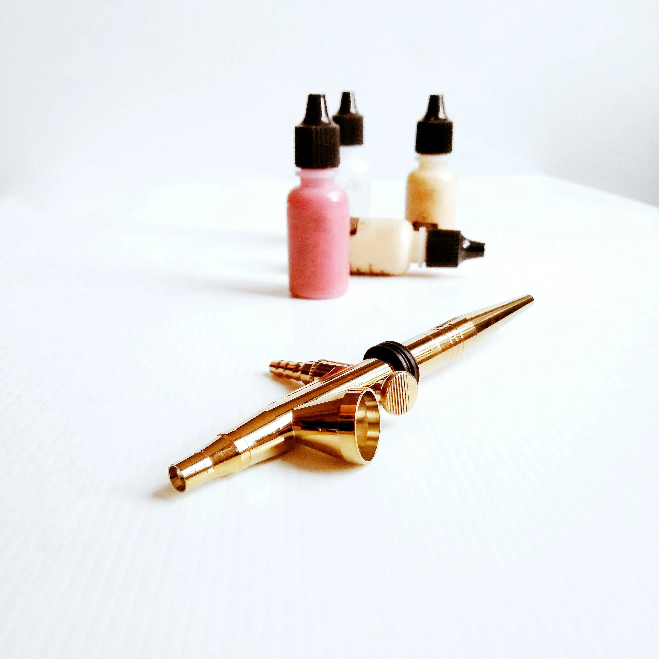 Gold spray tan gun with bottles of spray tan solution in pink, beige, and white in the background.