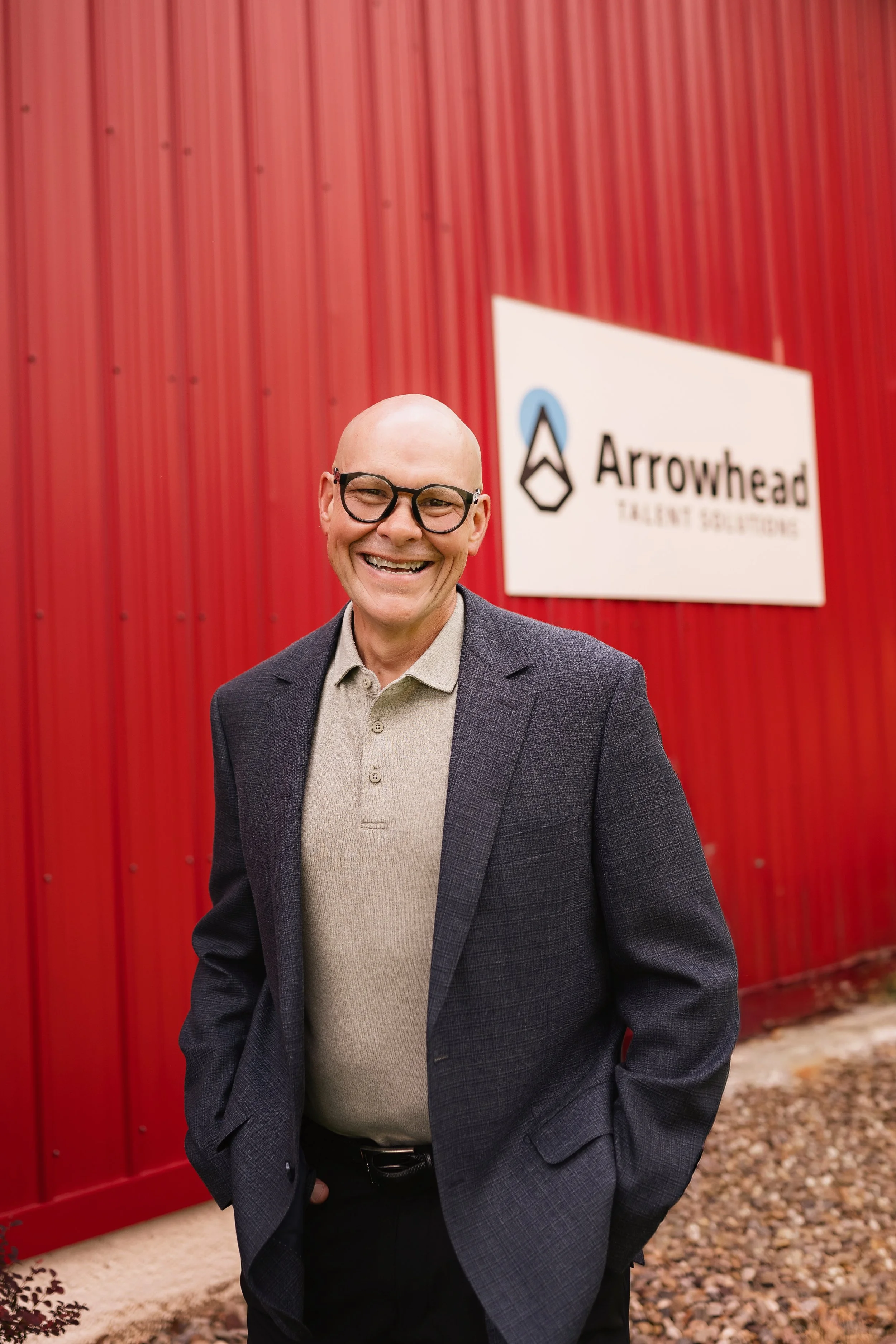 A smiling man with glasses and a bald head standing outdoors in front of a red building with a sign that reads "Arrowhead Talent Solutions".