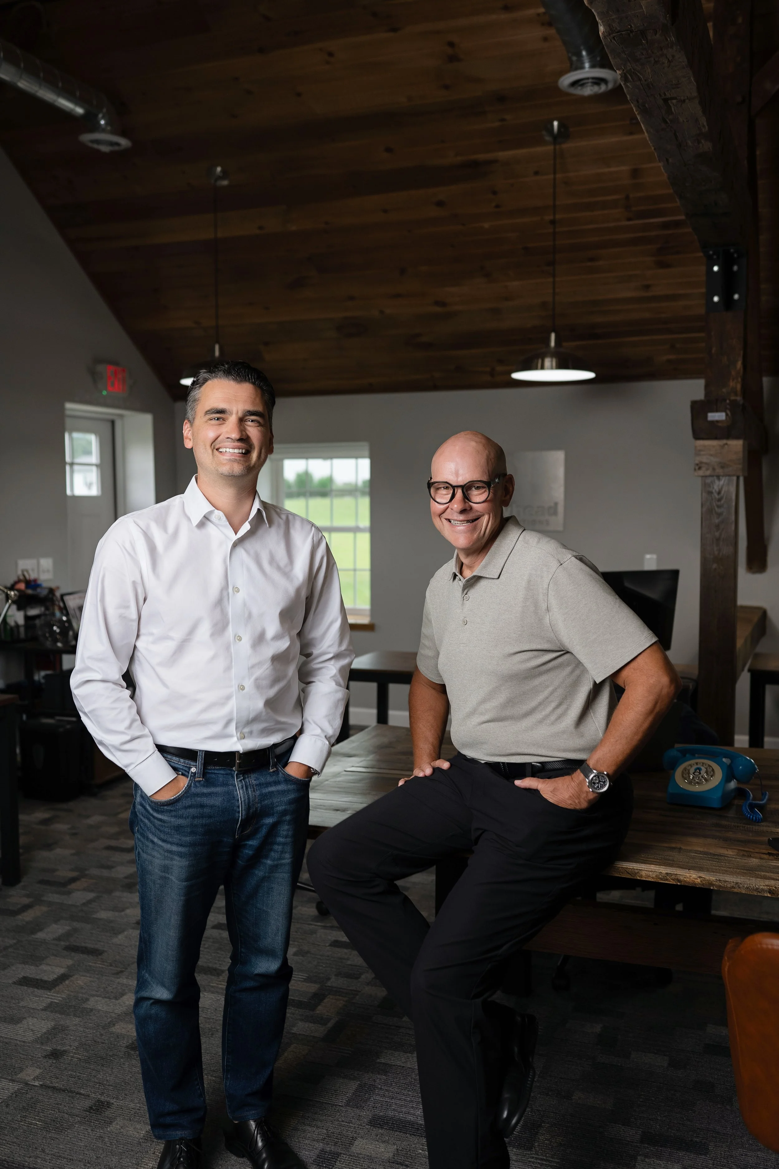 Two men in an office space, one standing and the other sitting on a desk, smiling at the camera. One man is wearing a white shirt and jeans, the other is wearing a beige polo shirt and black pants.