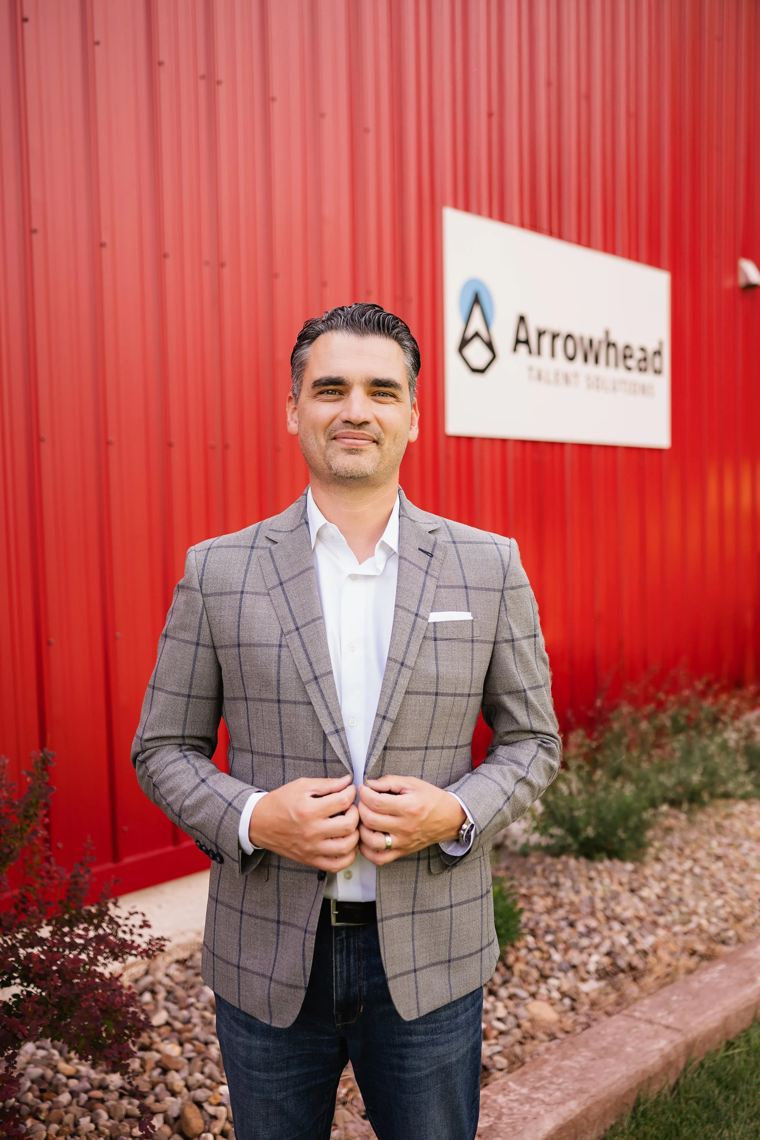 A man in a gray plaid blazer and white shirt standing outdoors in front of a red building with a sign that says 'Arrowhead Electric Solutions'.