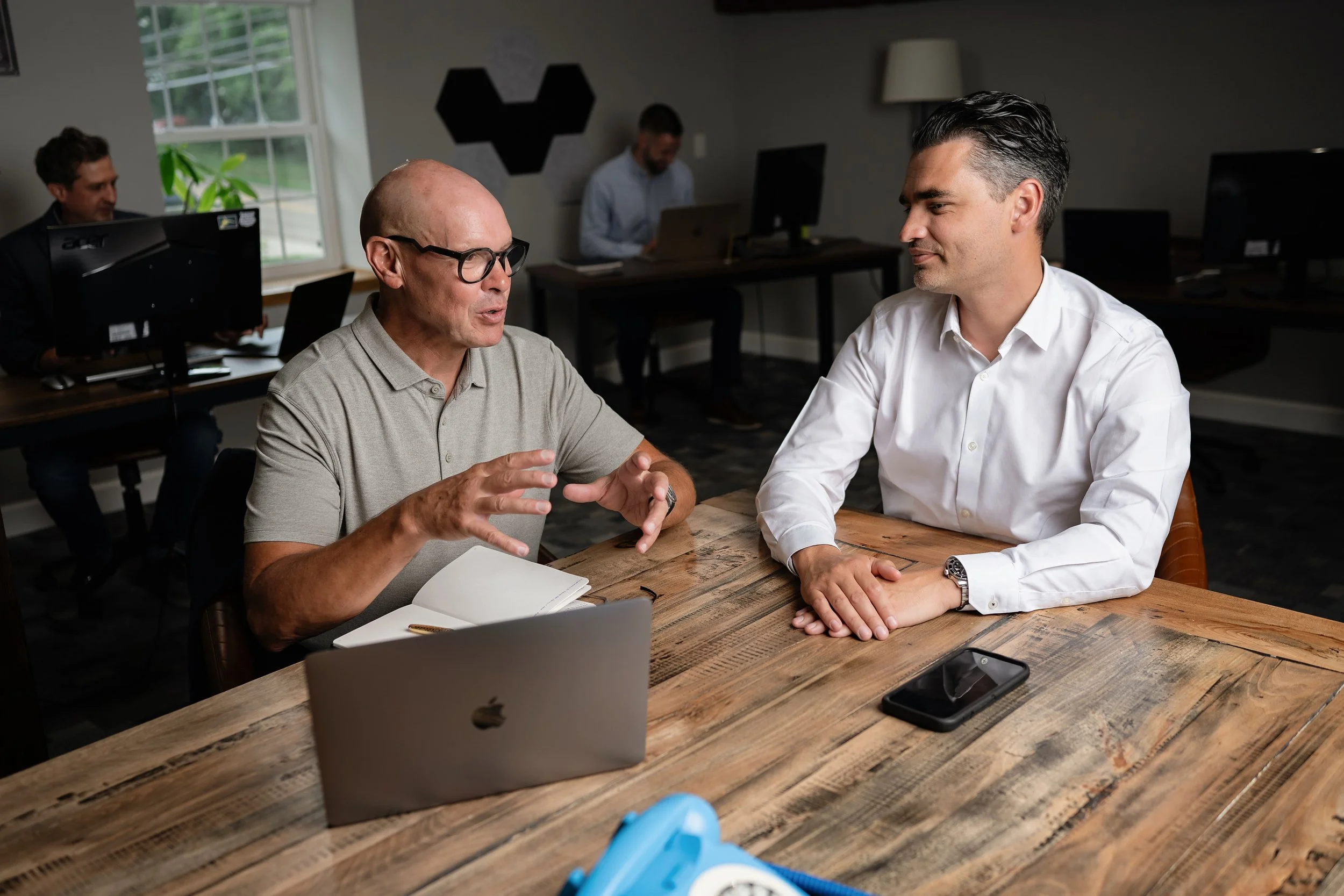 Two men sitting at a wooden table having a conversation in an office, with two other men working at desks in the background.