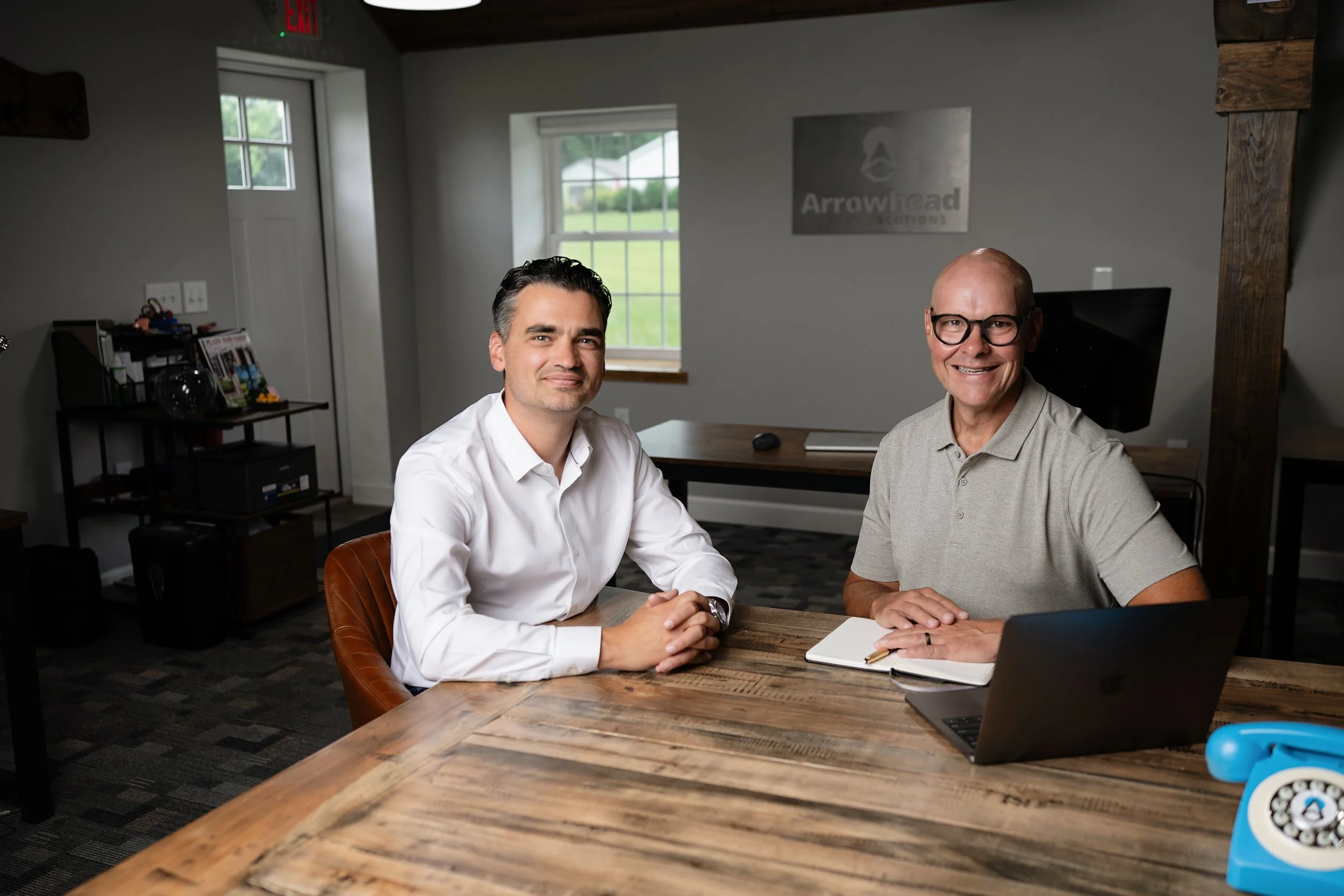 Two men sitting at a wooden table in an office, smiling at the camera. One man is wearing a white shirt, the other is wearing a gray polo. The office has a window, a computer, a notebook, and a sign on the wall that says 'Arrowhead.'
