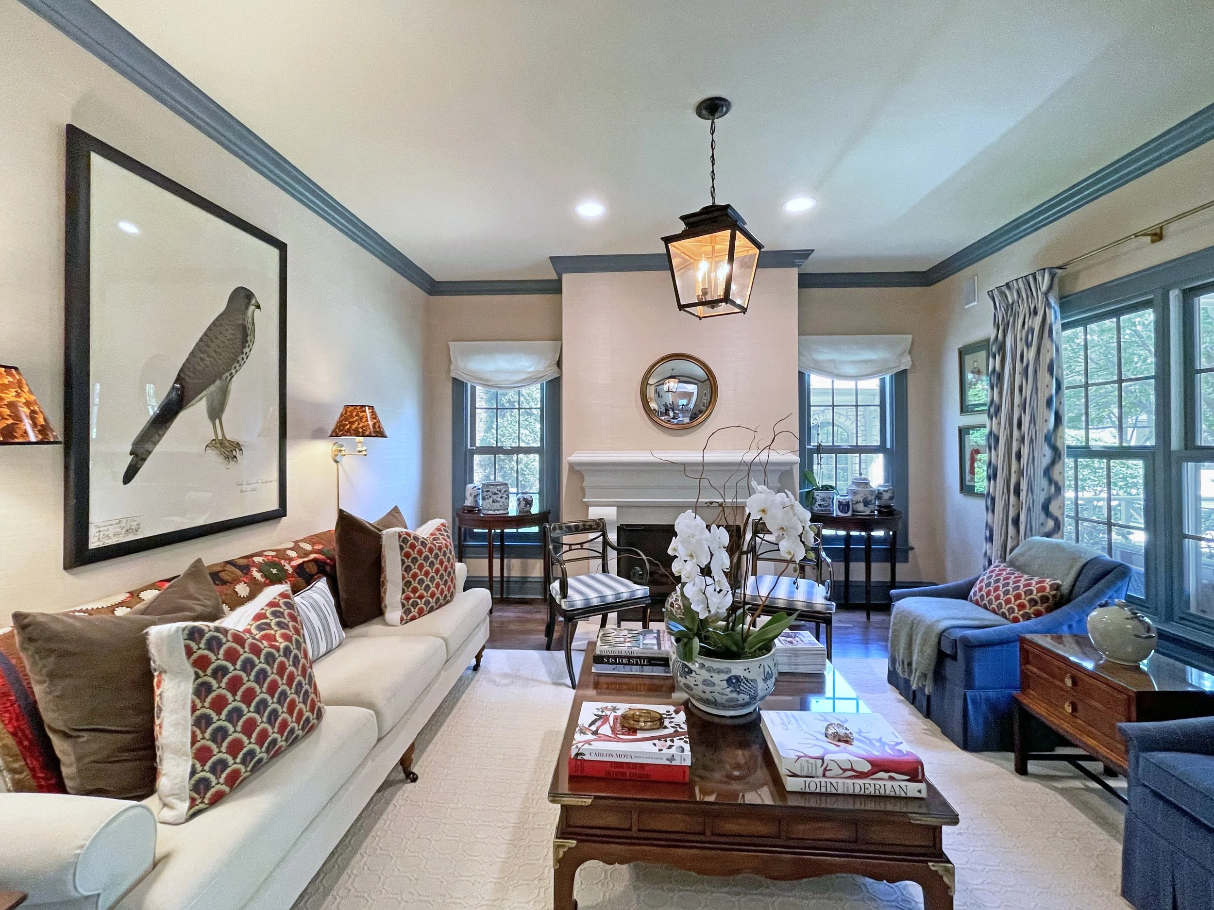 Living room with a white sofa adorned with patterned pillows, a glass coffee table with art books and a bowl of green moss balls, a fireplace with a round mirror above it, and a window with beige curtains.
