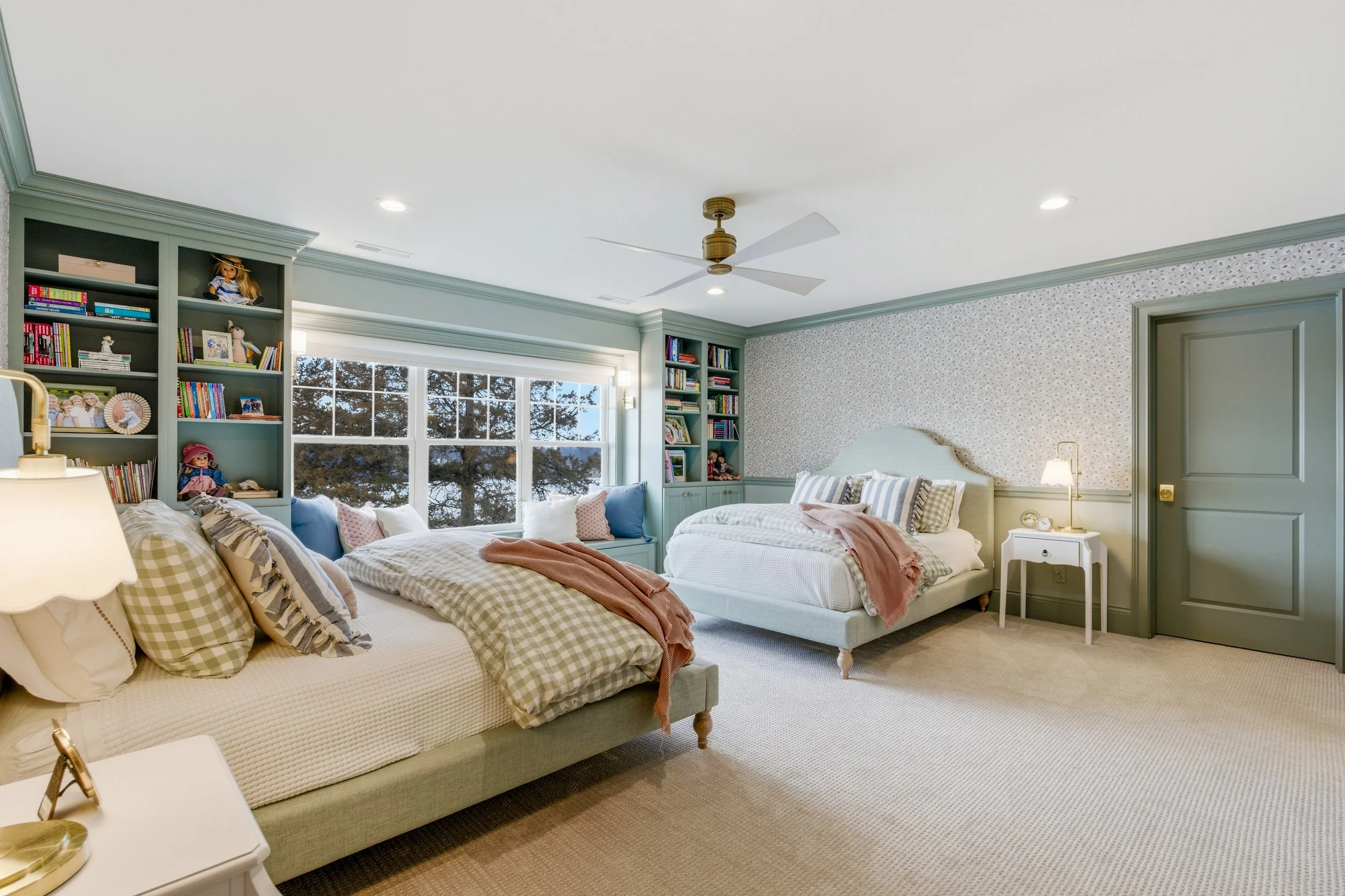 A formal dining room with a rectangular table set with dinnerware and a centerpiece, wooden chairs with white fabric bows, a chandelier with candle-like lights, floral wallpaper, a window with striped curtains, and a sideboard with lamps and decorative items.
