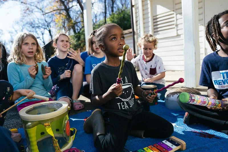 Group of children sitting outside on a blue carpet, playing musical instruments and enjoying a sunny day.