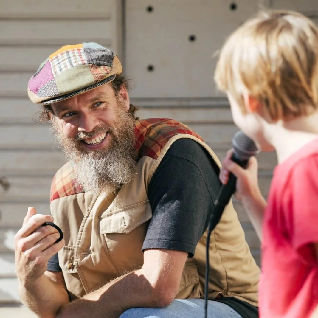 A man with a beard, wearing a colorful patchwork cap and a beige vest, smiling at a young child with blonde hair, who is holding a microphone.