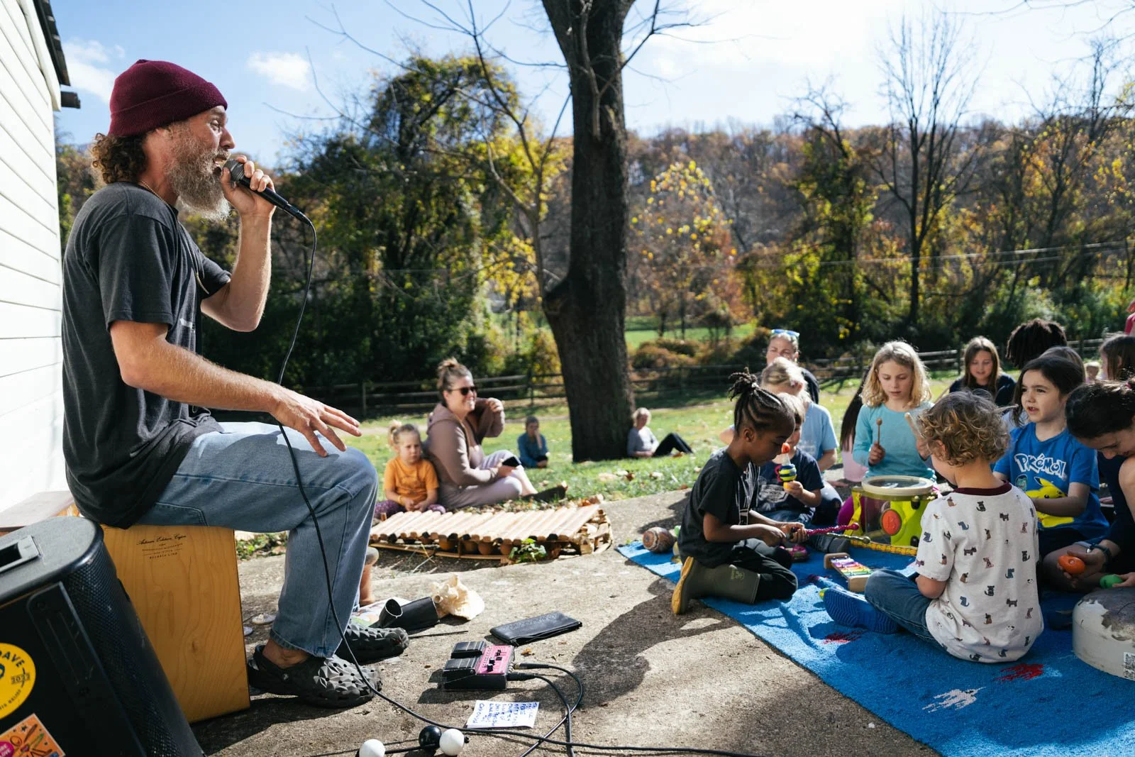 A man with a beard and curly hair wearing a maroon beanie sits outdoors playing a harmonica for children. Kids sit on a blue outdoor carpet, playing with rainsticks and percussion instruments. Others sit on the grass, observing, with a large tree in the background, during a sunny fall day.