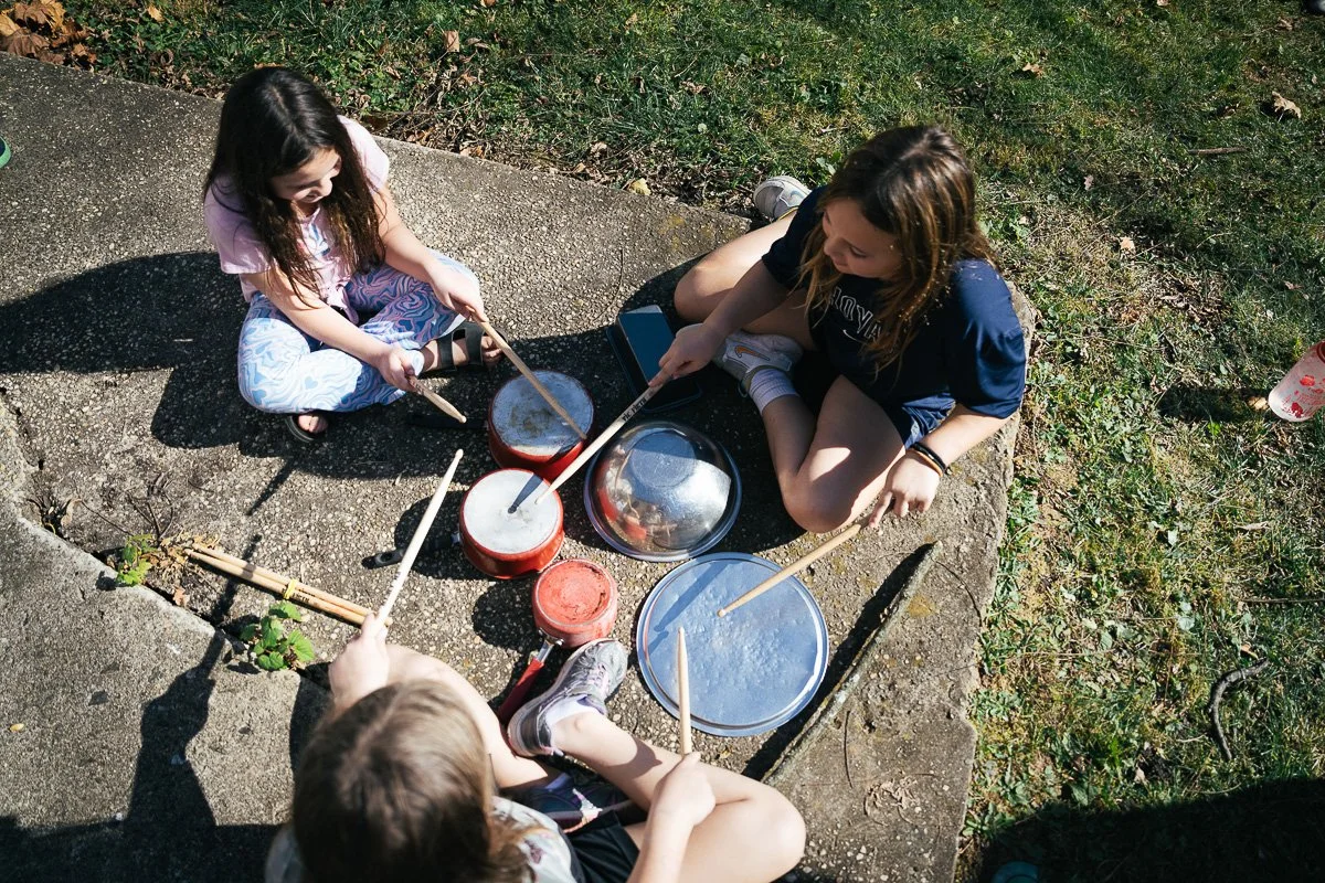 Three children sitting on a sidewalk playing drums and percussion instruments outdoors on a sunny day.