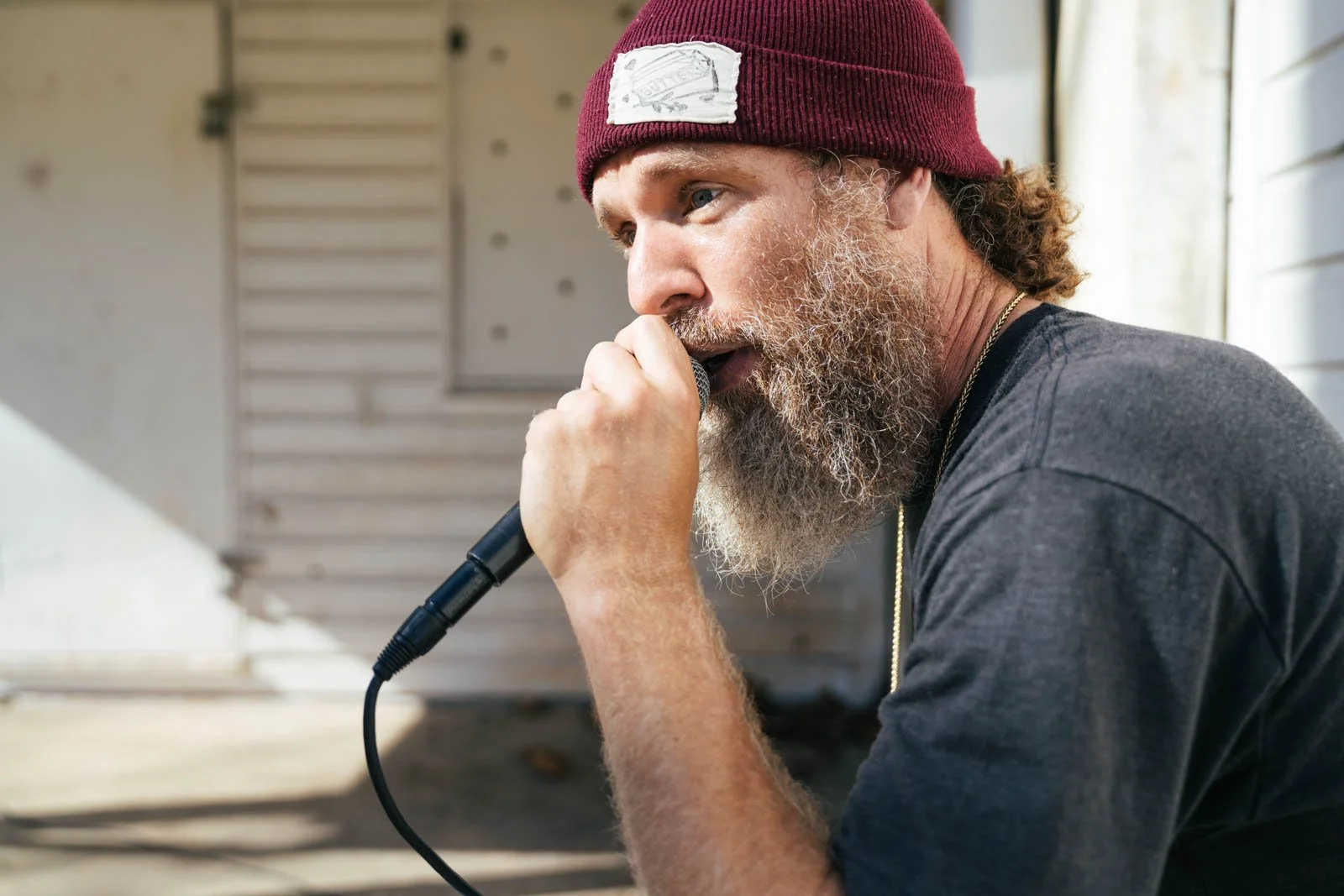 A man with a gray beard and curly hair wearing a burgundy beanie and a dark t-shirt, singing into a microphone indoors near a window.