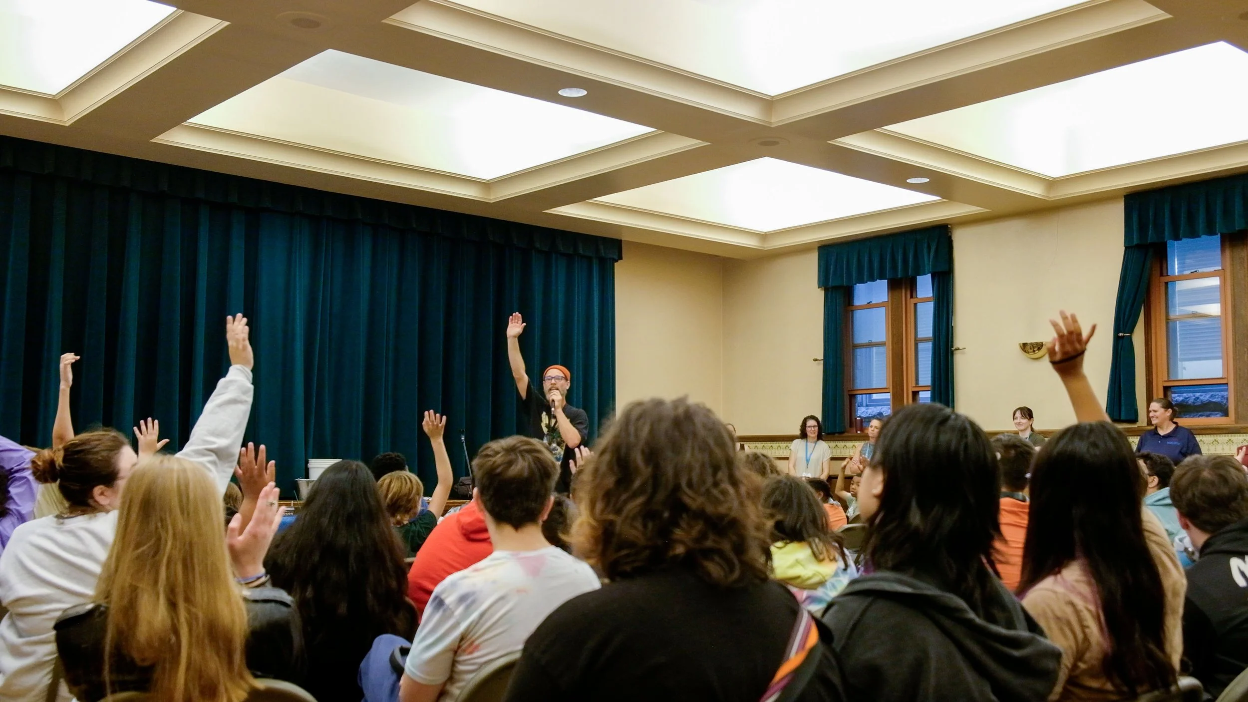 A man wearing glasses and an orange beanie speaking into a microphone with a raised hand in a conference room filled with seated people, some of whom are raising their hands.