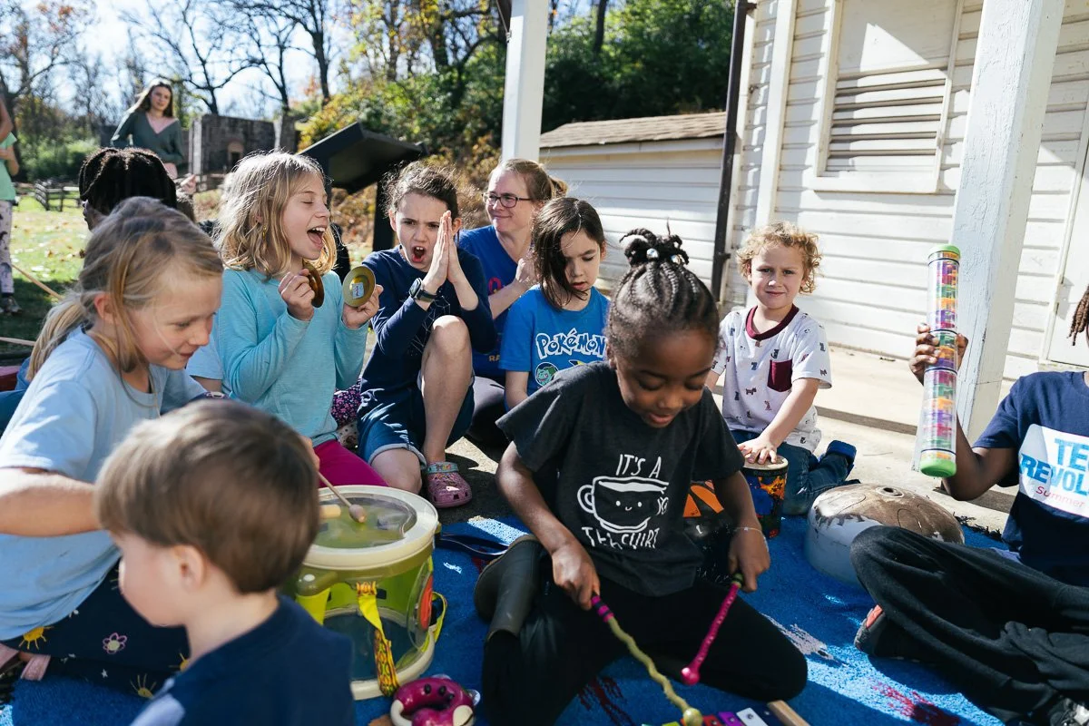 Children and adults sitting on a blue blanket outdoors, engaging in musical activities with a drum, xylophone, and water bottles, during a sunny day.