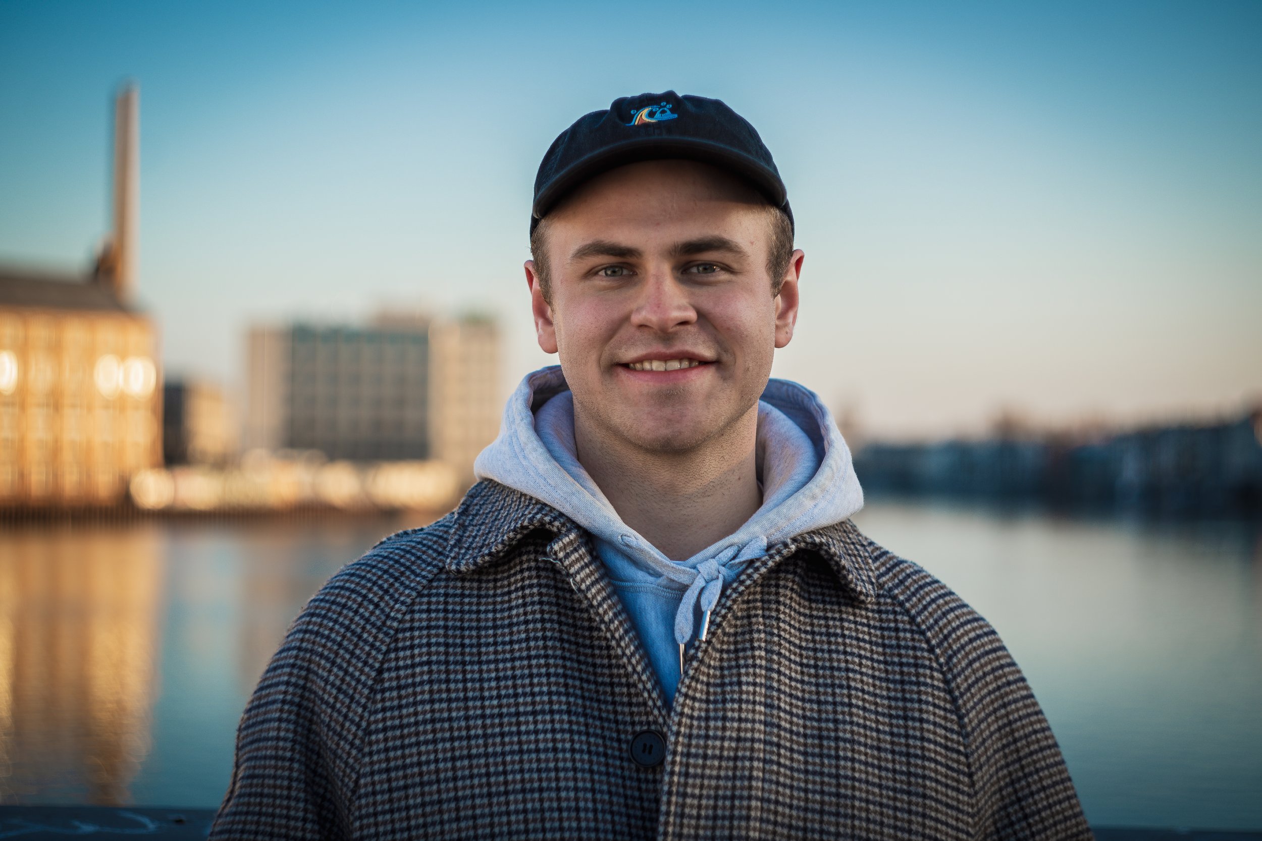 Young man smiling outdoors near a waterway with blurred buildings in the background, wearing a black cap and a checkered jacket.