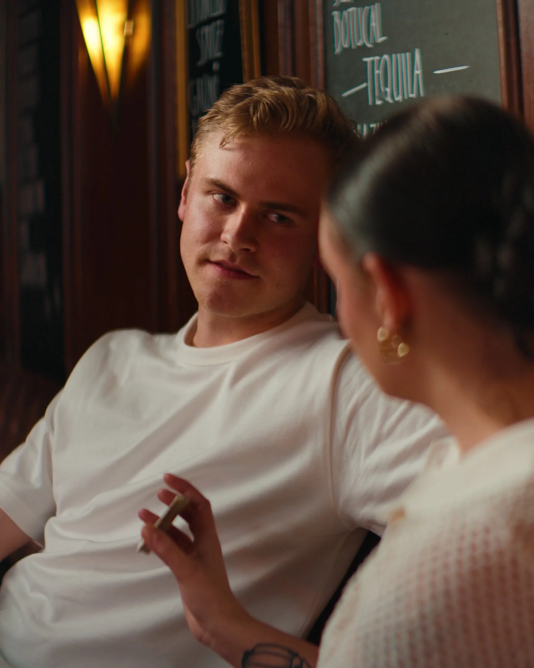 A young man and woman sitting at a table in a dimly lit restaurant or bar, engaged in conversation. The man is looking at the woman with a serious expression, and the woman is holding a cigarette.