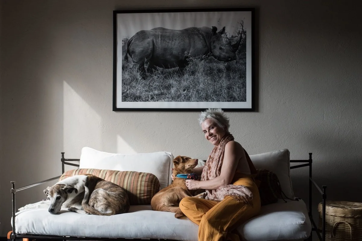 An elderly woman with short white hair sitting on a white couch with two dogs, one resting and one sitting next to her. A large black and white photograph of a rhinoceros hangs on the wall behind them.