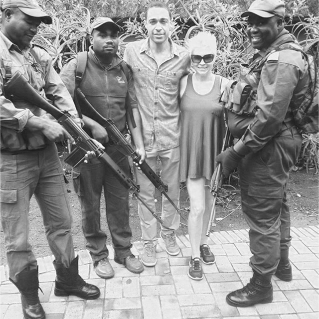 Group of four armed soldiers and two civilians smiling outdoors in front of greenery.