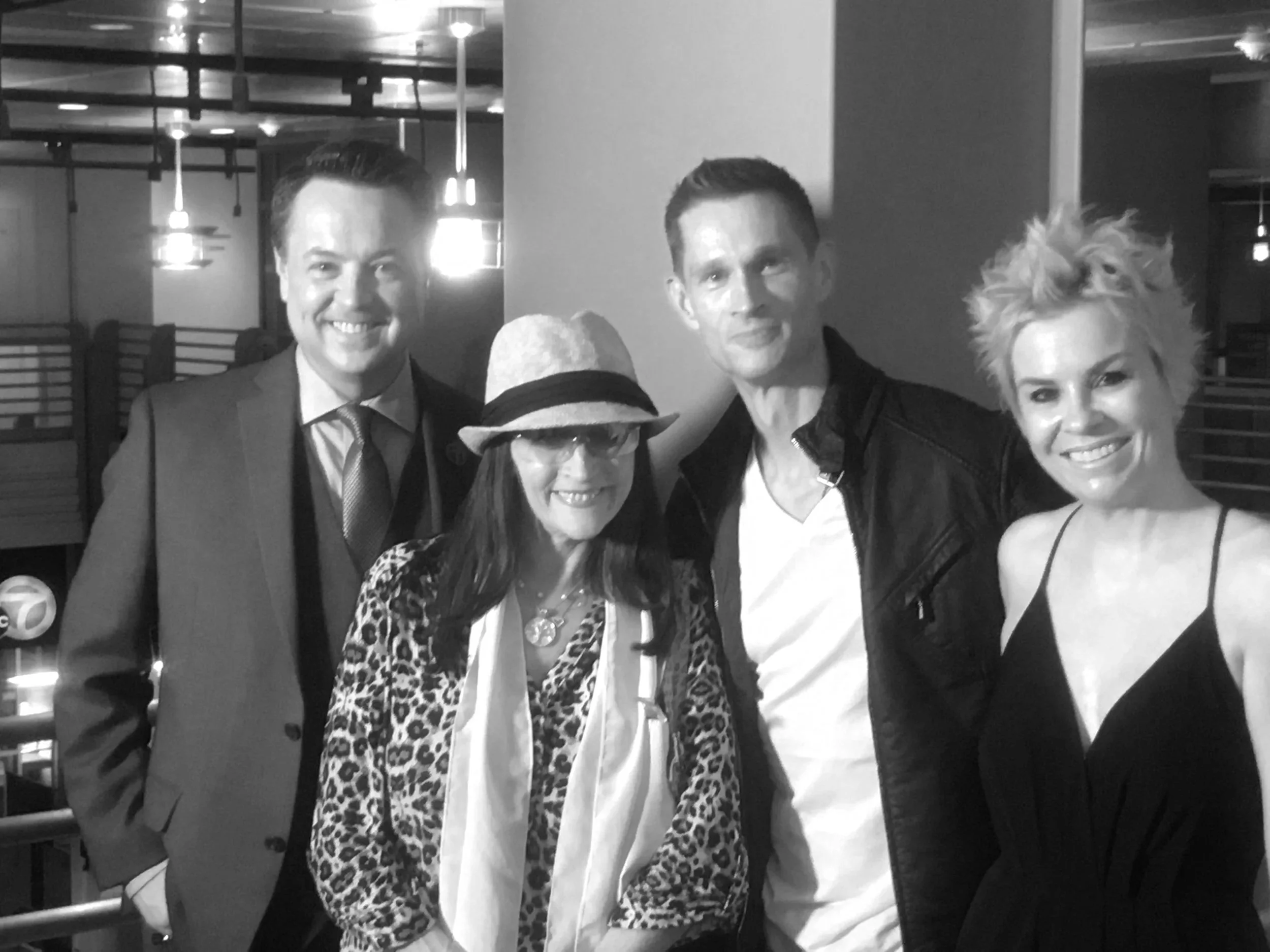 Group of four people smiling, dressed in formal and casual clothing, standing inside a restaurant or bar with modern decor.