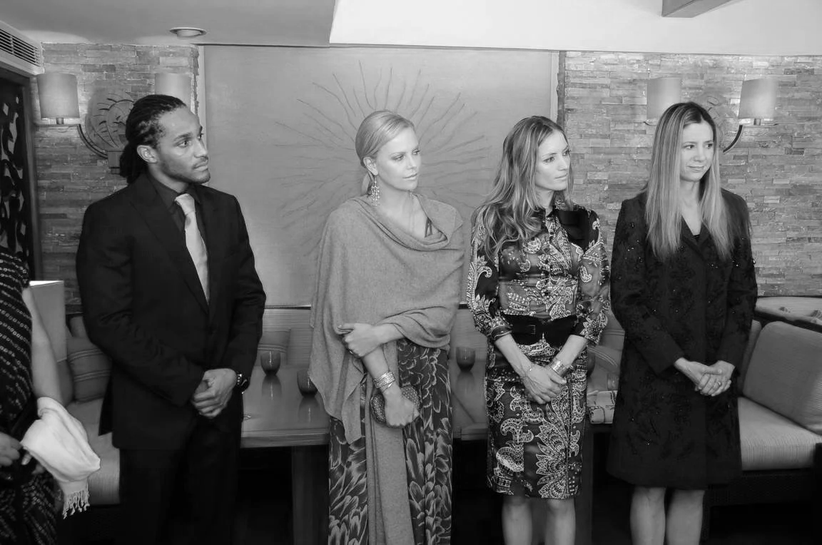 Four people standing indoors, dressed formally, in front of a wall with a decorative design and wall-mounted lights.