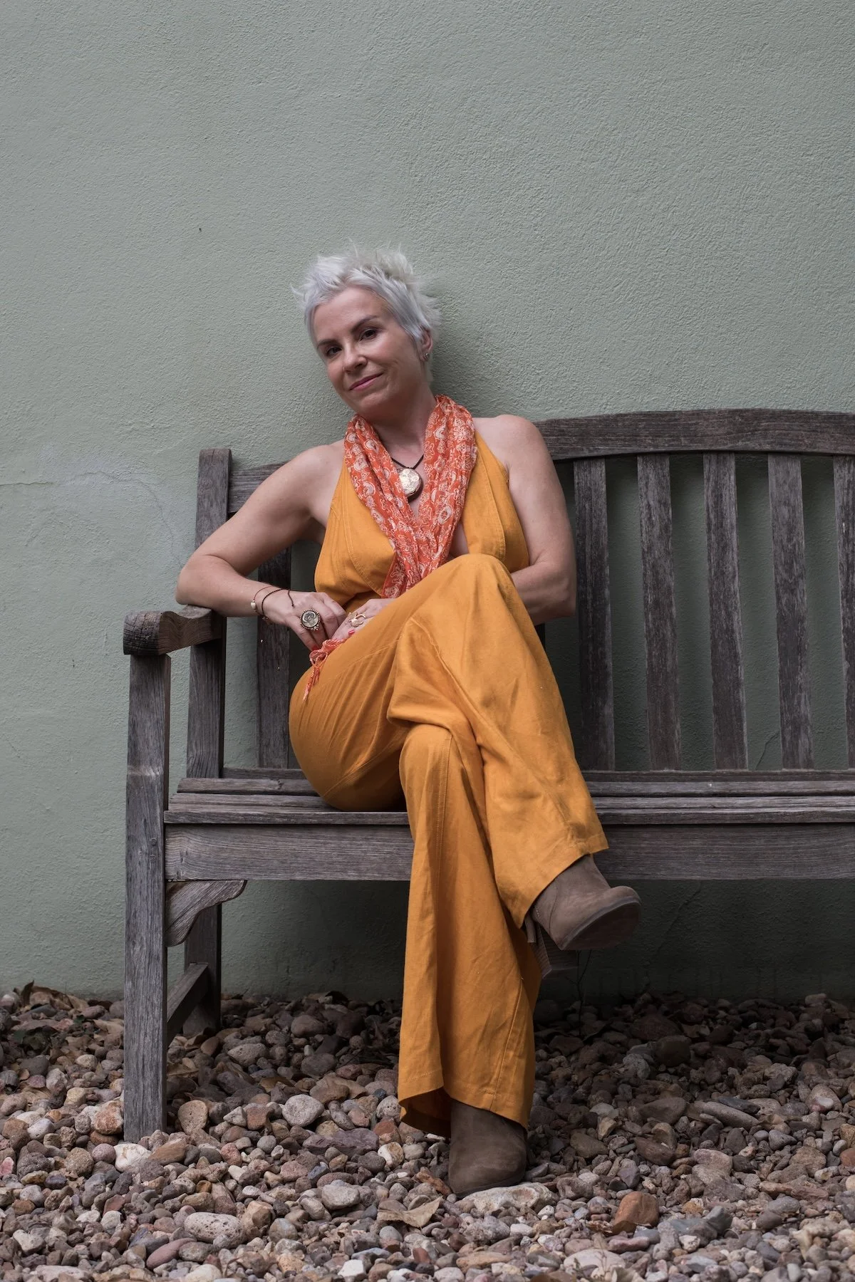 A woman with short, white hair sitting on a wooden bench against a plain, light green wall. She is wearing a mustard yellow sleeveless jumpsuit, a red patterned scarf, a large pendant necklace, and brown boots. She has one leg crossed over the other and is looking confidently at the camera.
