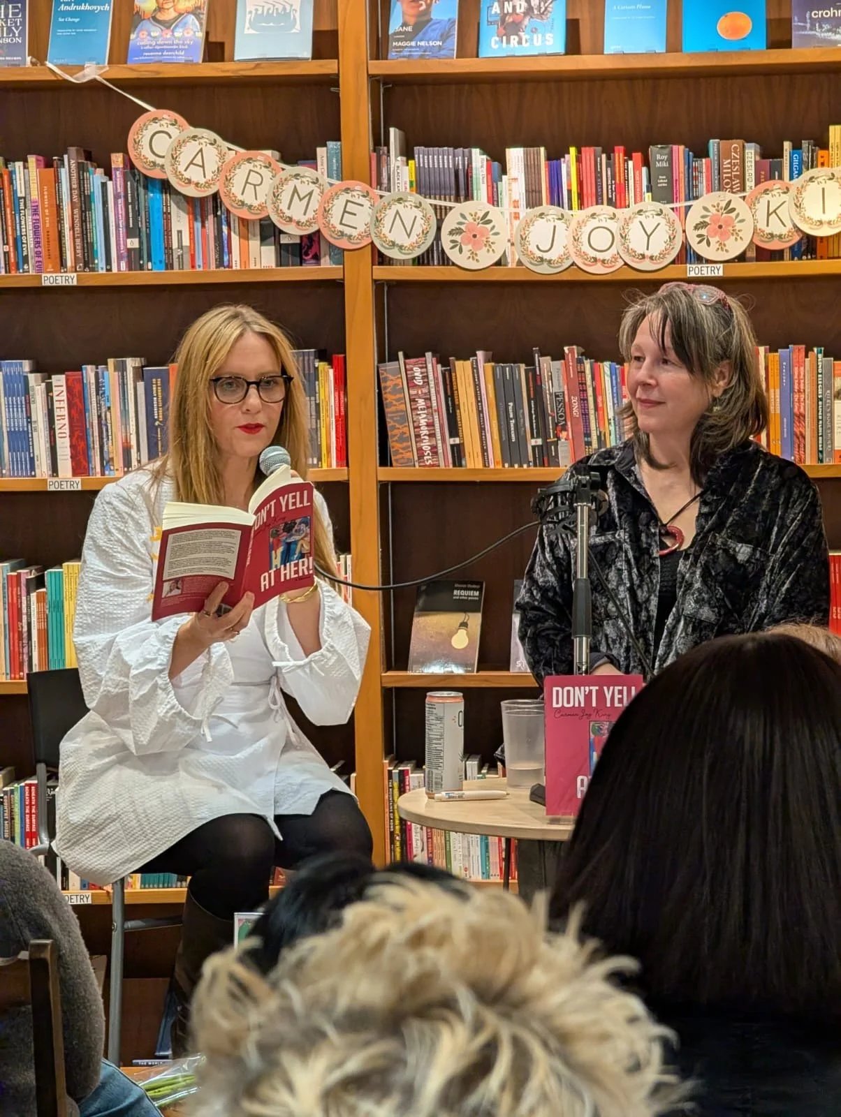 Two women sitting in a bookstore or library during a book reading event. One woman is speaking into a microphone and holding a book titled 'Don't Yell at Her,' while the other woman is sitting beside her. A decorative banner above them reads 'Sometimes Joy.' There are bookshelves filled with books behind them and some audience members visible in the foreground.