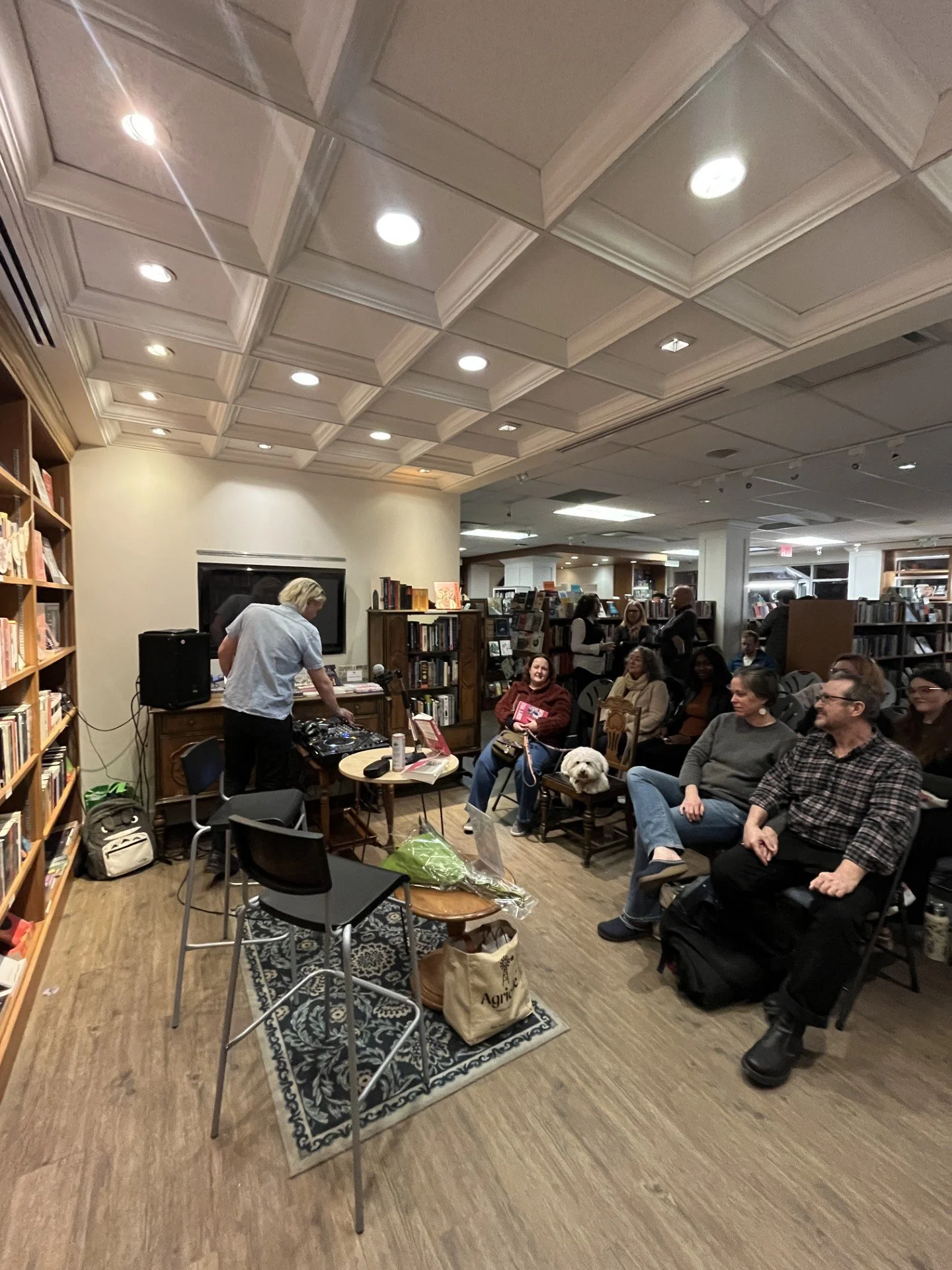 People attending a book reading or presentation at a bookstore or library. A DJ or sound technician is managing equipment at the front, with a small dog sitting on a woman's lap among the audience.