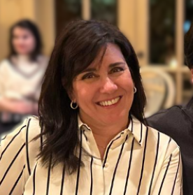 A woman smiling with dark hair, wearing a striped shirt, seated indoors with blurred people in the background.