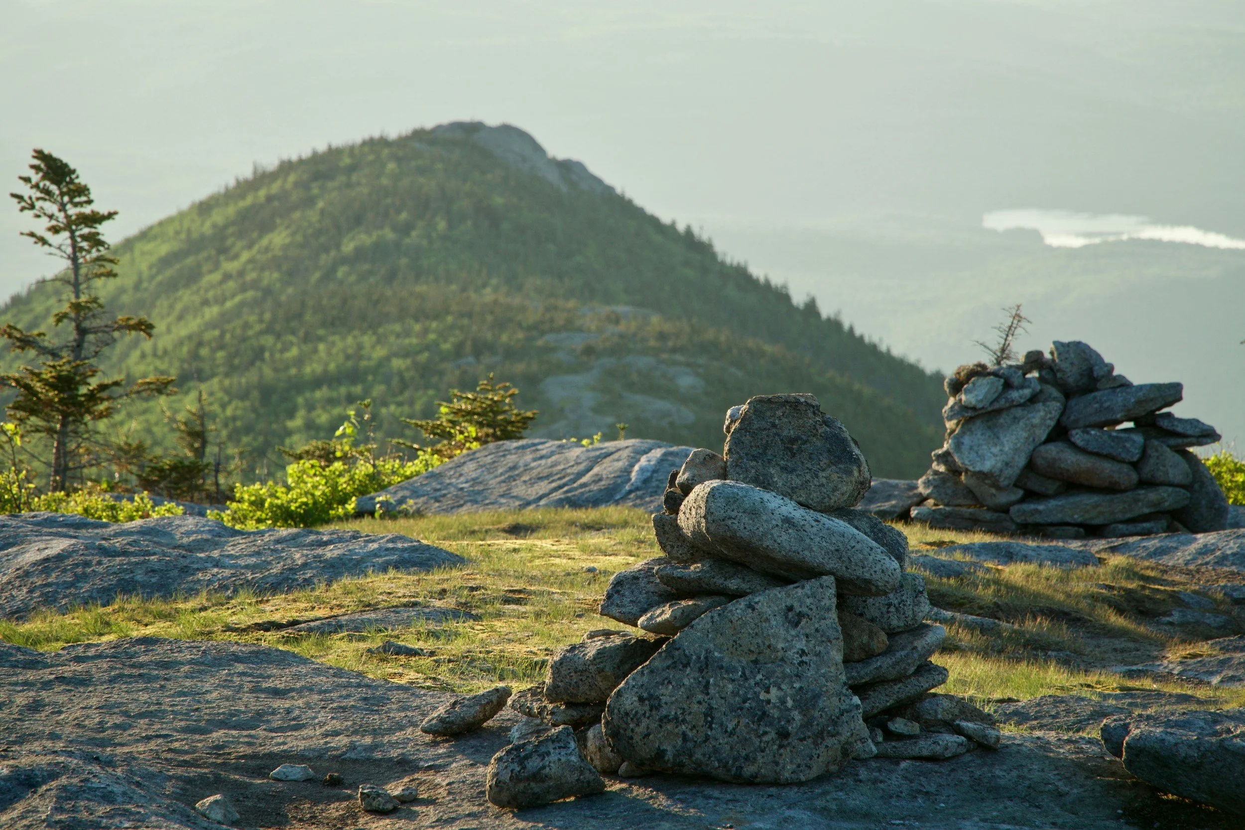 Two stacked cairns on rocky terrain with a mountain and green trees in the background during daylight.