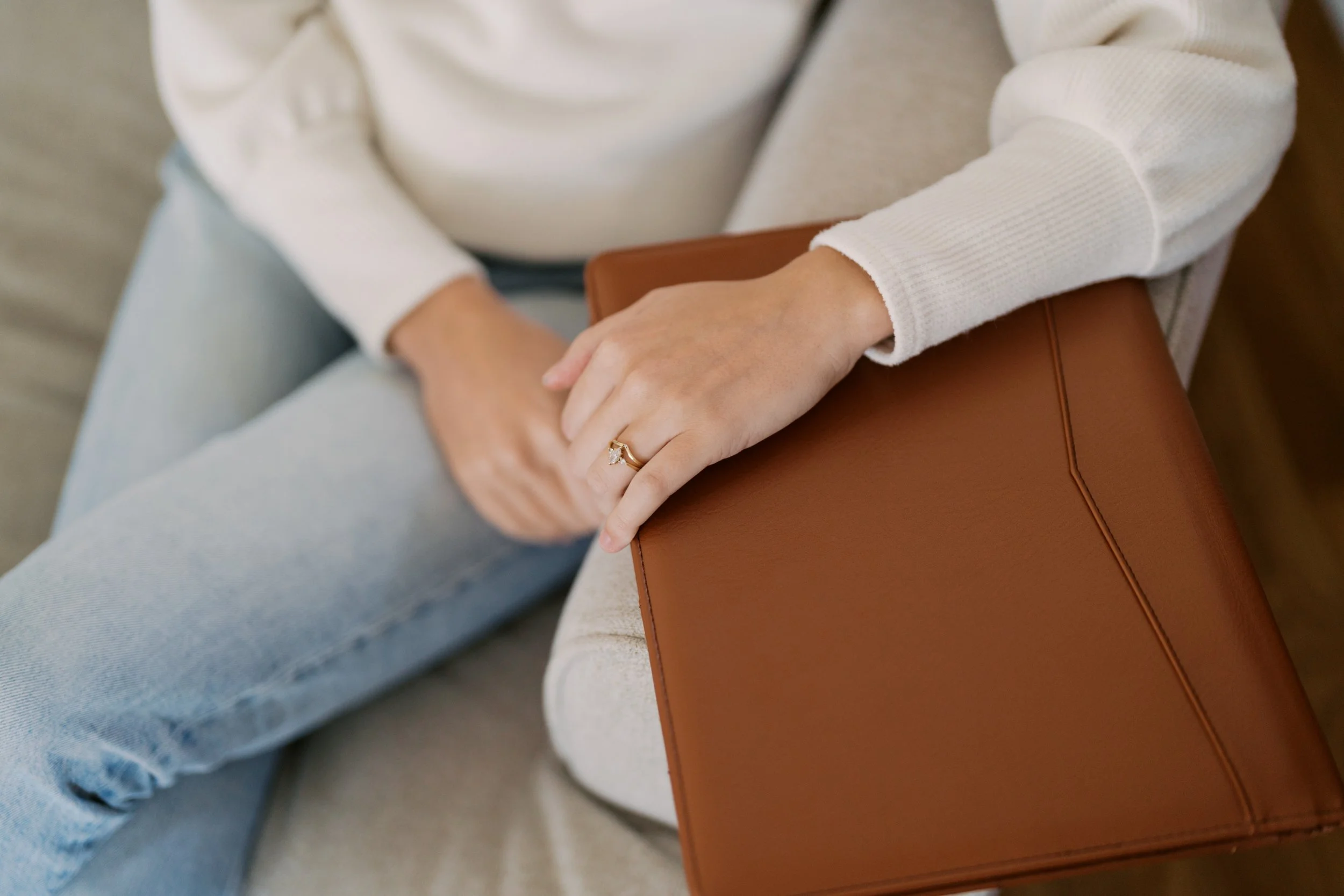 A person sitting on a light-colored chair, wearing a cream-colored sweater and light blue jeans, holding a brown leather bag on their lap with their left hand, which has a gold ring with a diamond.
