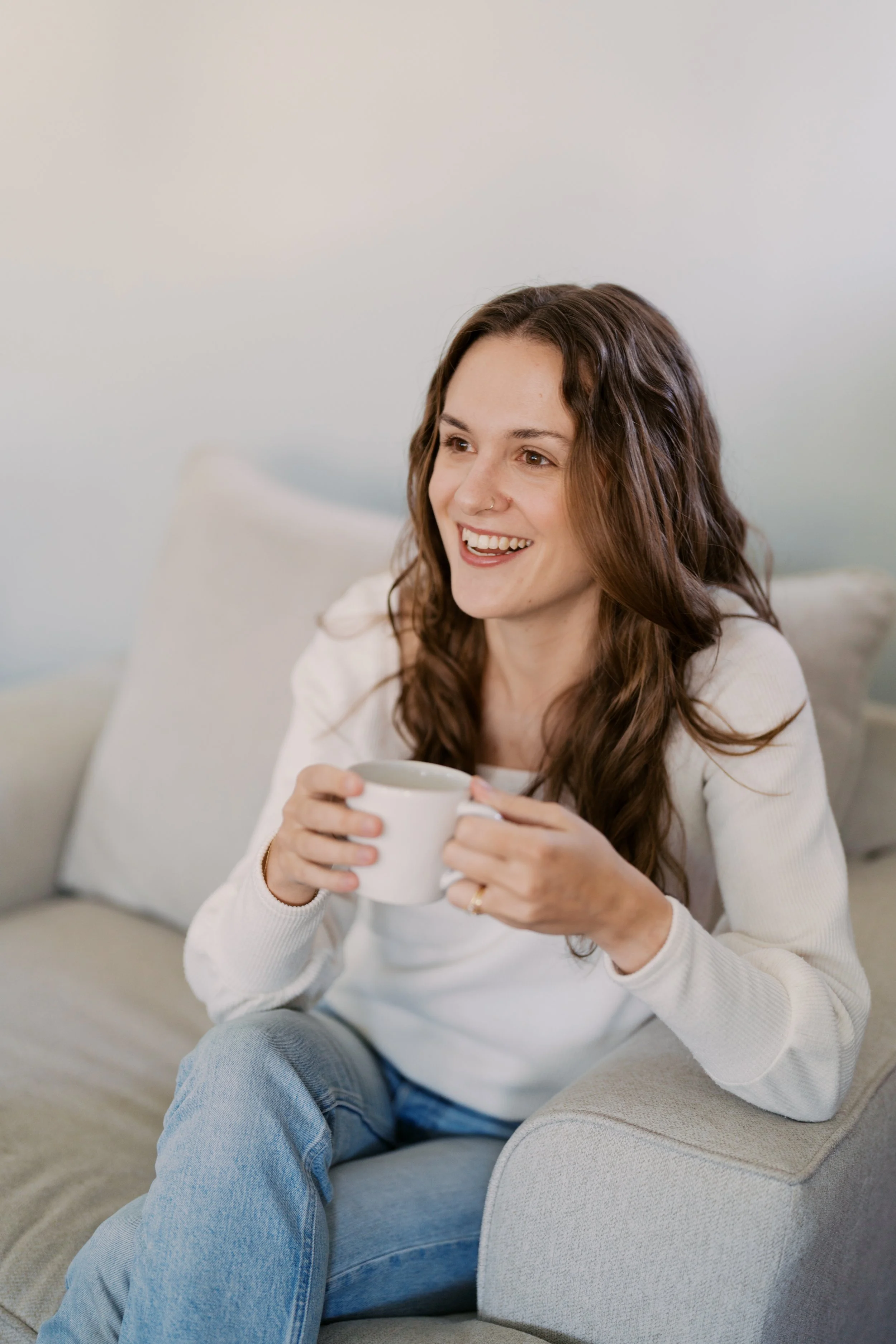A woman with long brown hair and a nose piercing sitting on a beige couch, smiling while holding a white mug.
