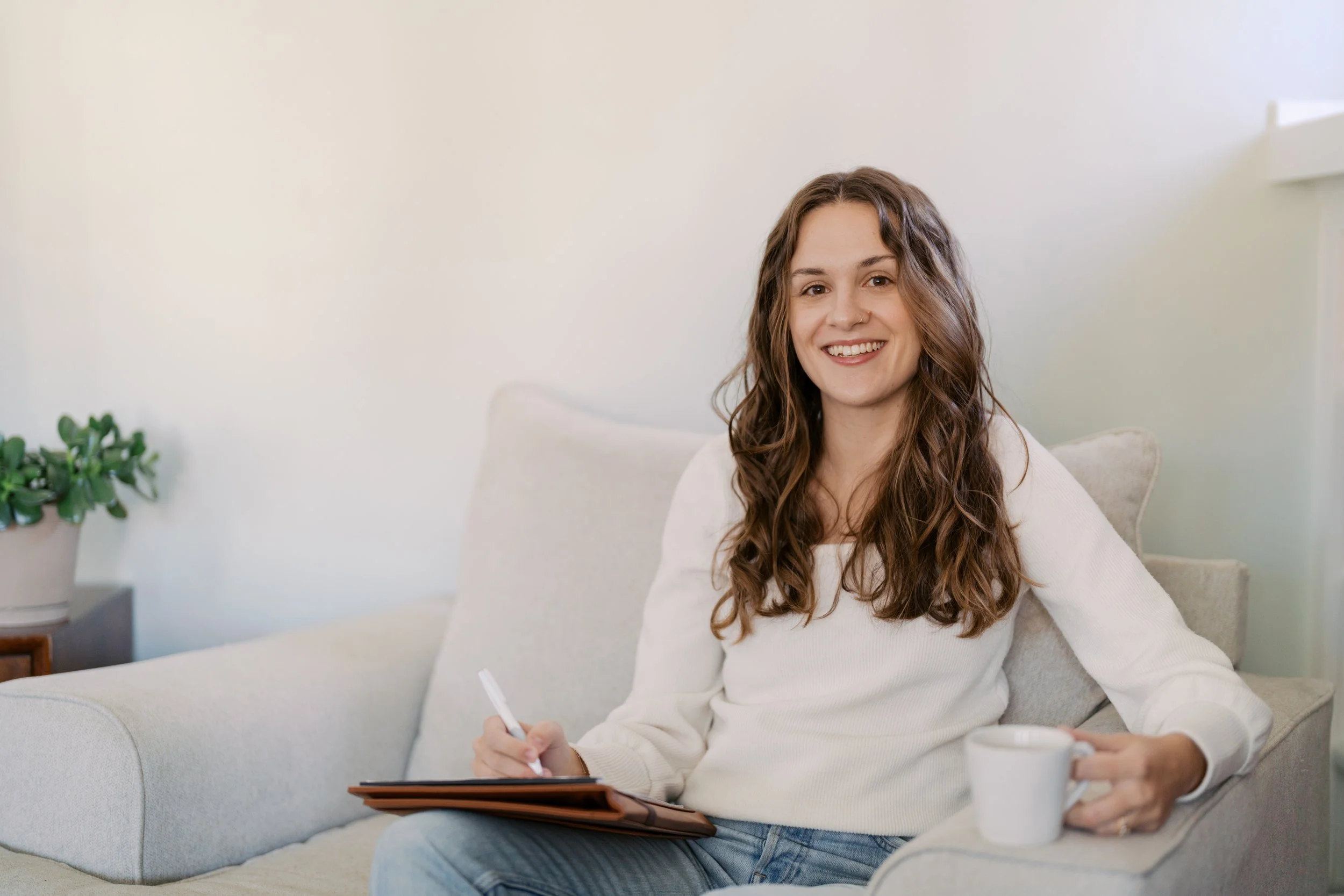 A smiling woman with long, wavy brown hair sitting on a beige couch holding a white mug in her left hand and a notebook with a pen in her right hand. She is wearing a cream-colored sweater and blue jeans in a cozy and well-lit room.