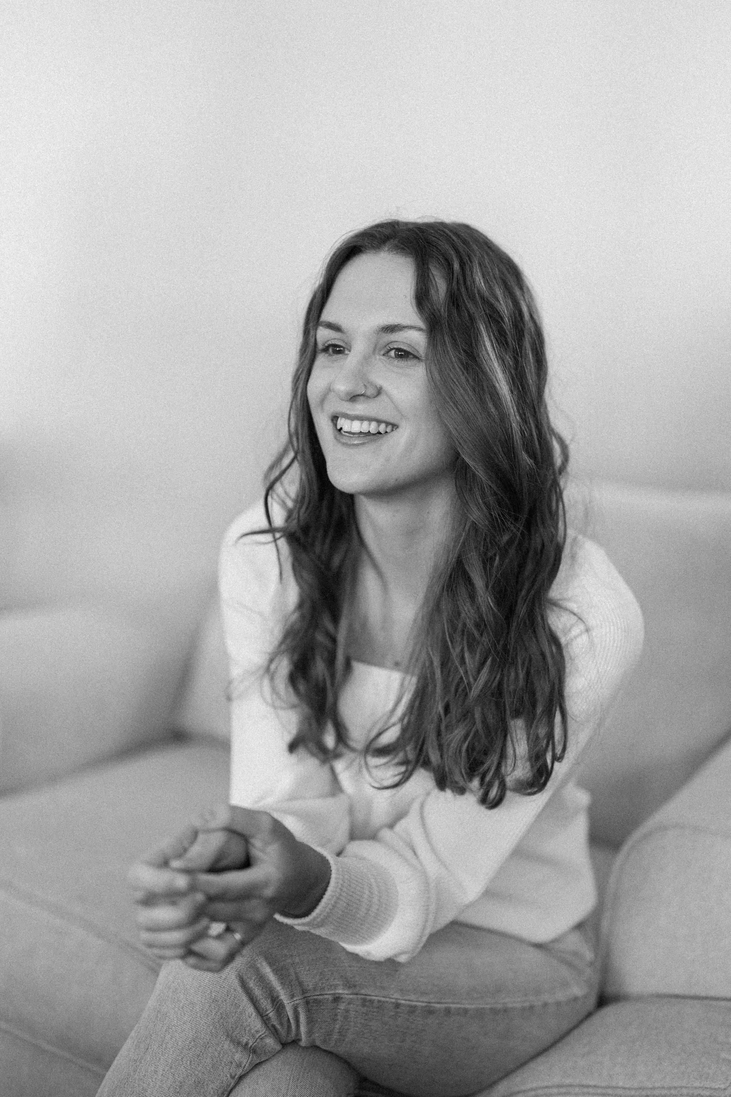 Black and white photo of a smiling woman with long wavy hair sitting on a couch, wearing a light-colored sweater and jeans, with her hands clasped together.