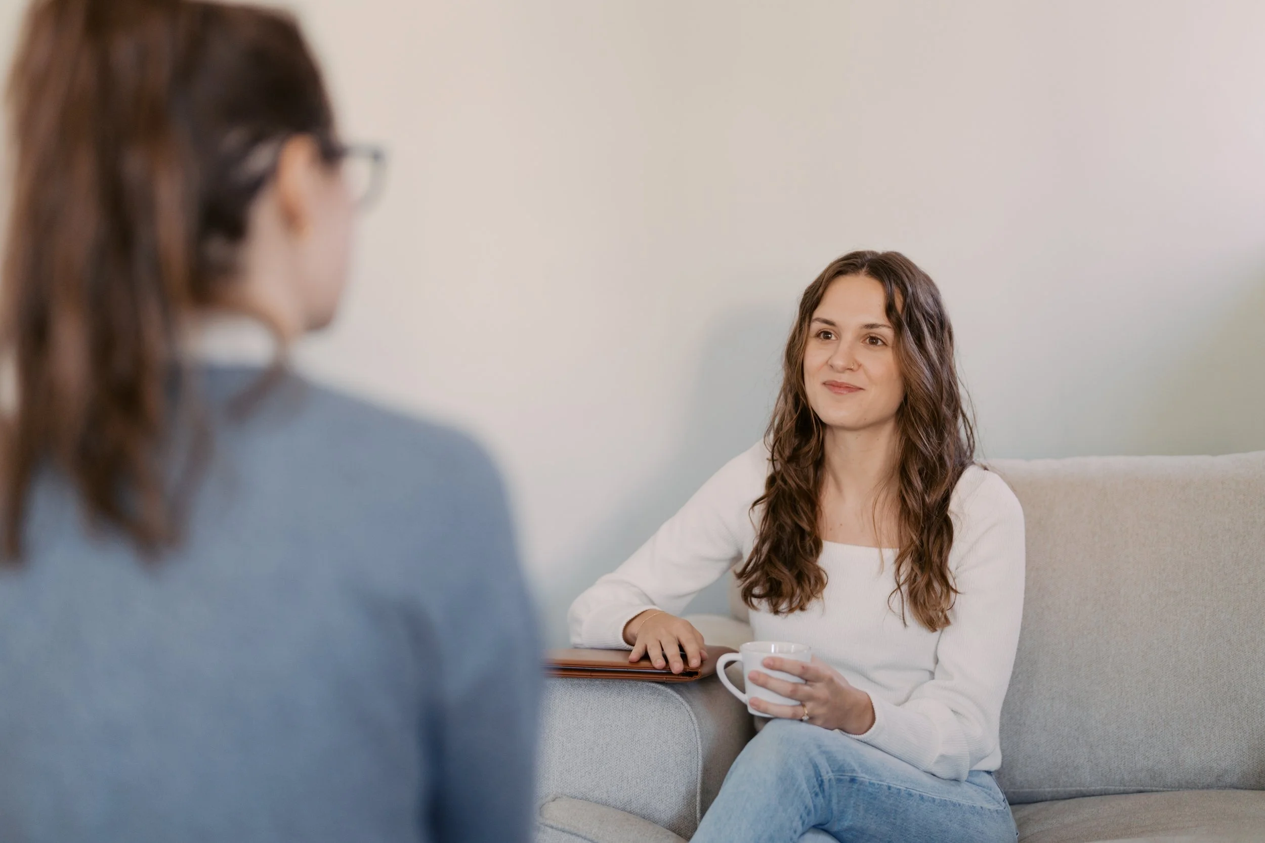 Woman sitting on a beige couch holding a white mug, talking to another woman with glasses, in a living room with plain white wall.