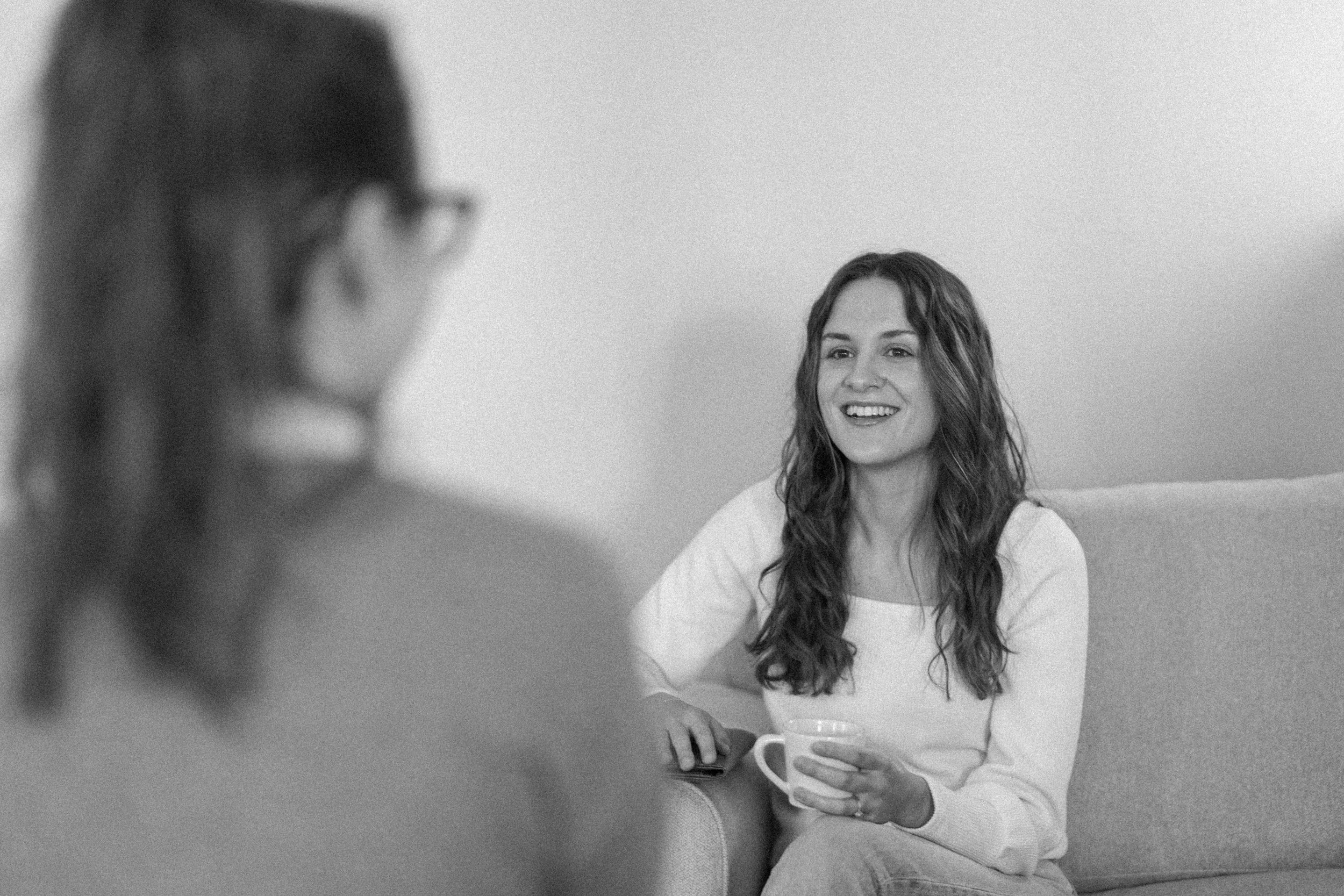 Two women sitting on a couch having a conversation, one is smiling and holding a cup, the other is out of focus.