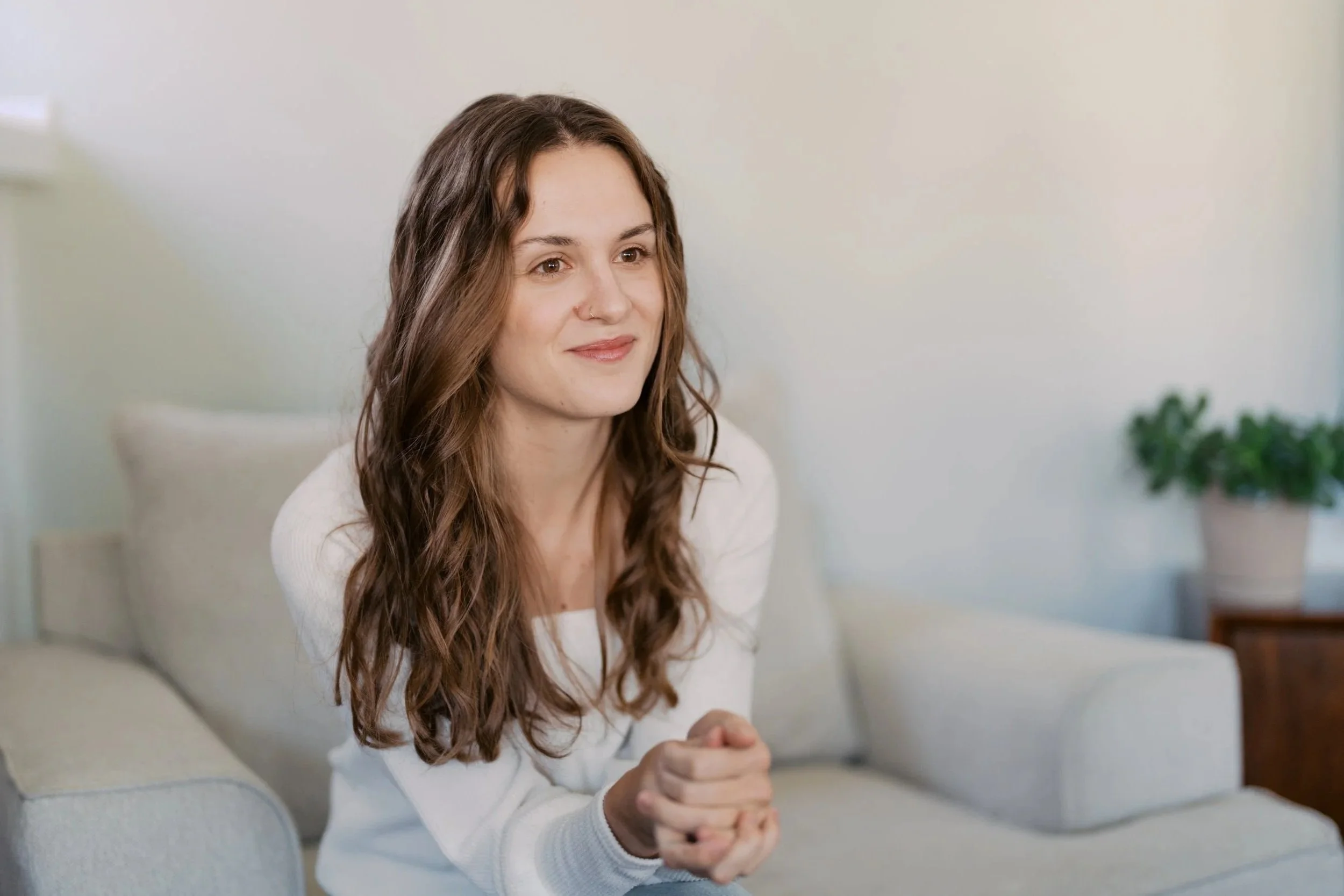 A young woman with long, wavy brown hair sitting on a beige sofa in a well-lit room, smiling softly.
