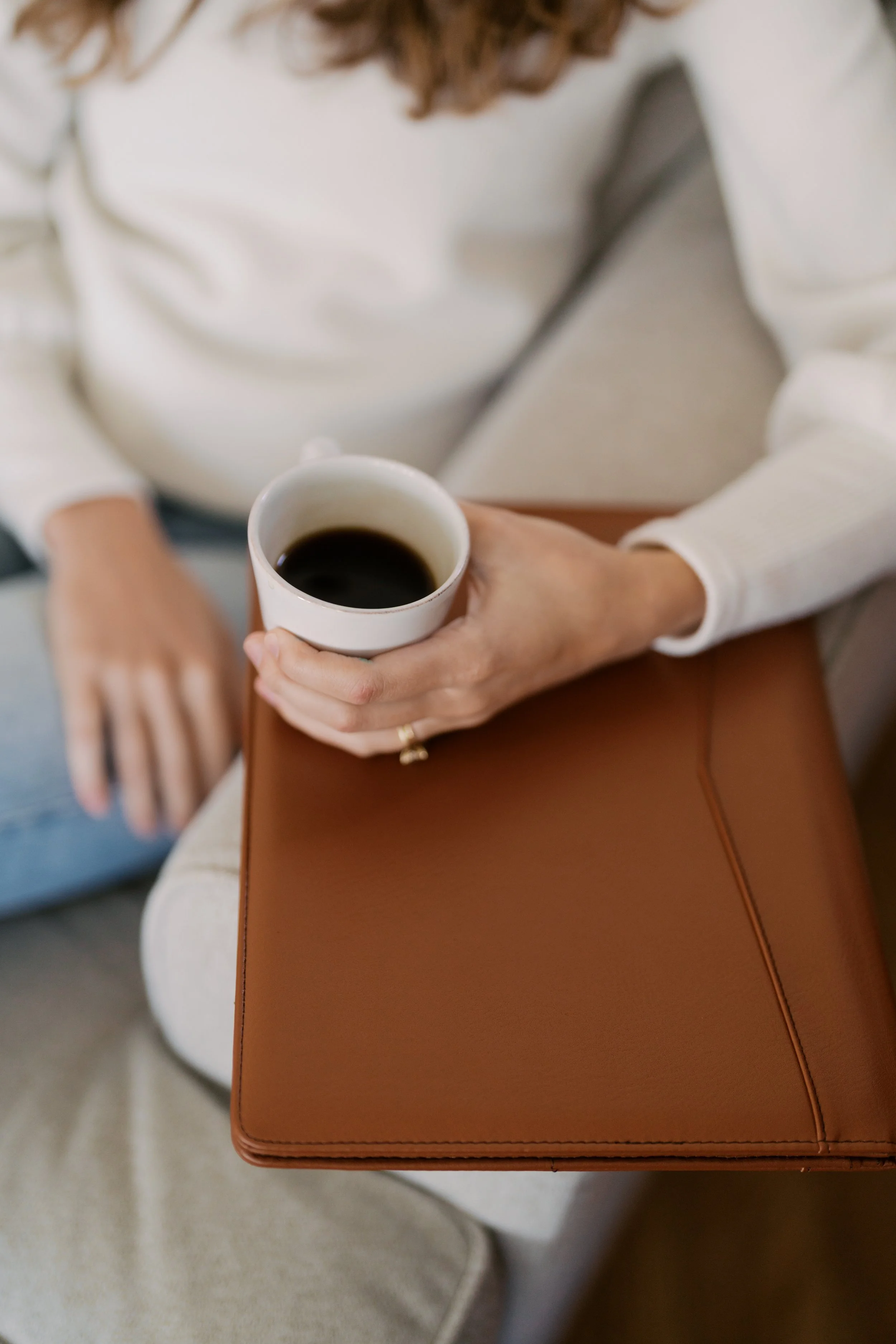 Person holding a white cup of black coffee while sitting on a sofa with a brown leather bag on lap.