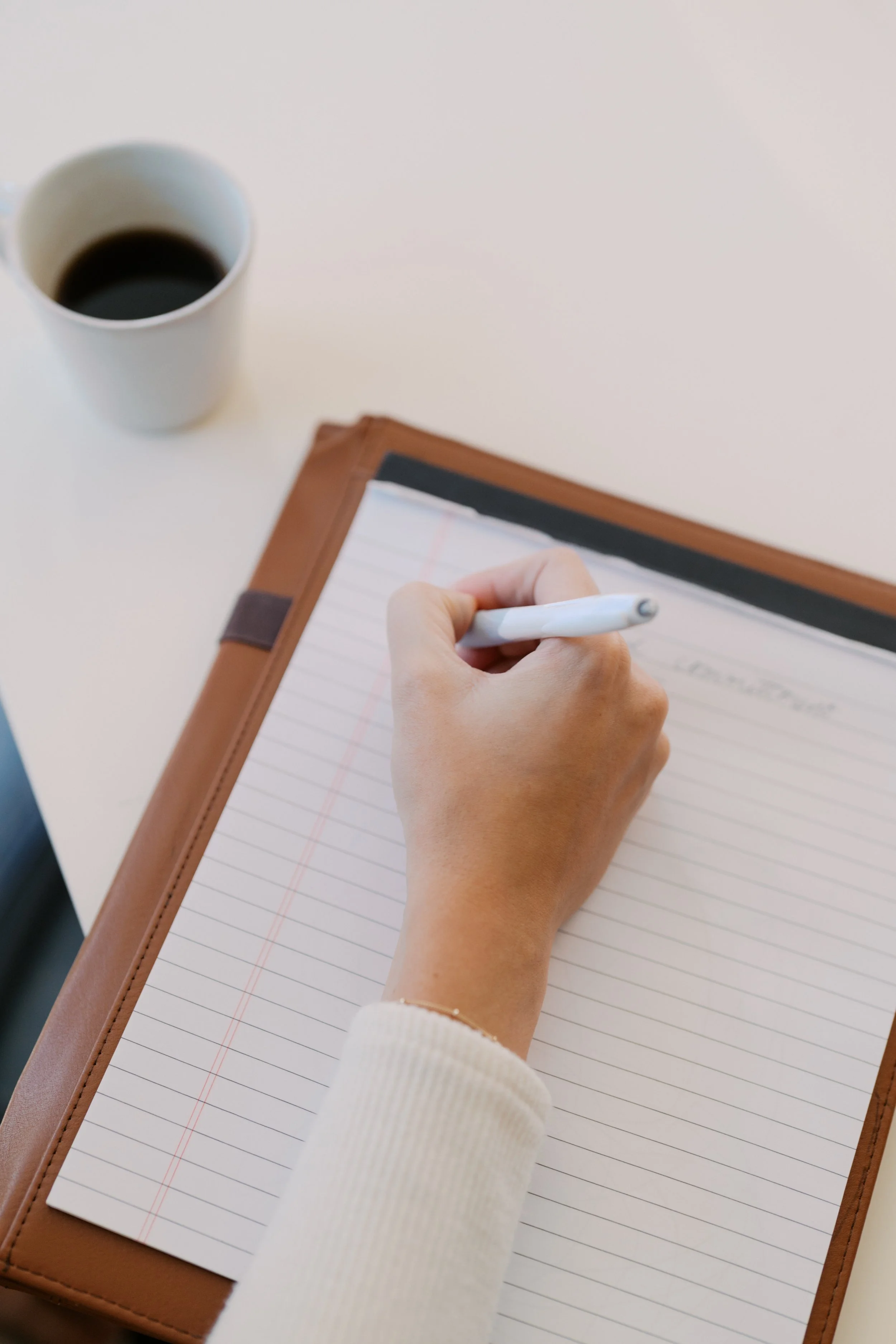 A person writing in a notebook with a cup of coffee nearby on a white table.