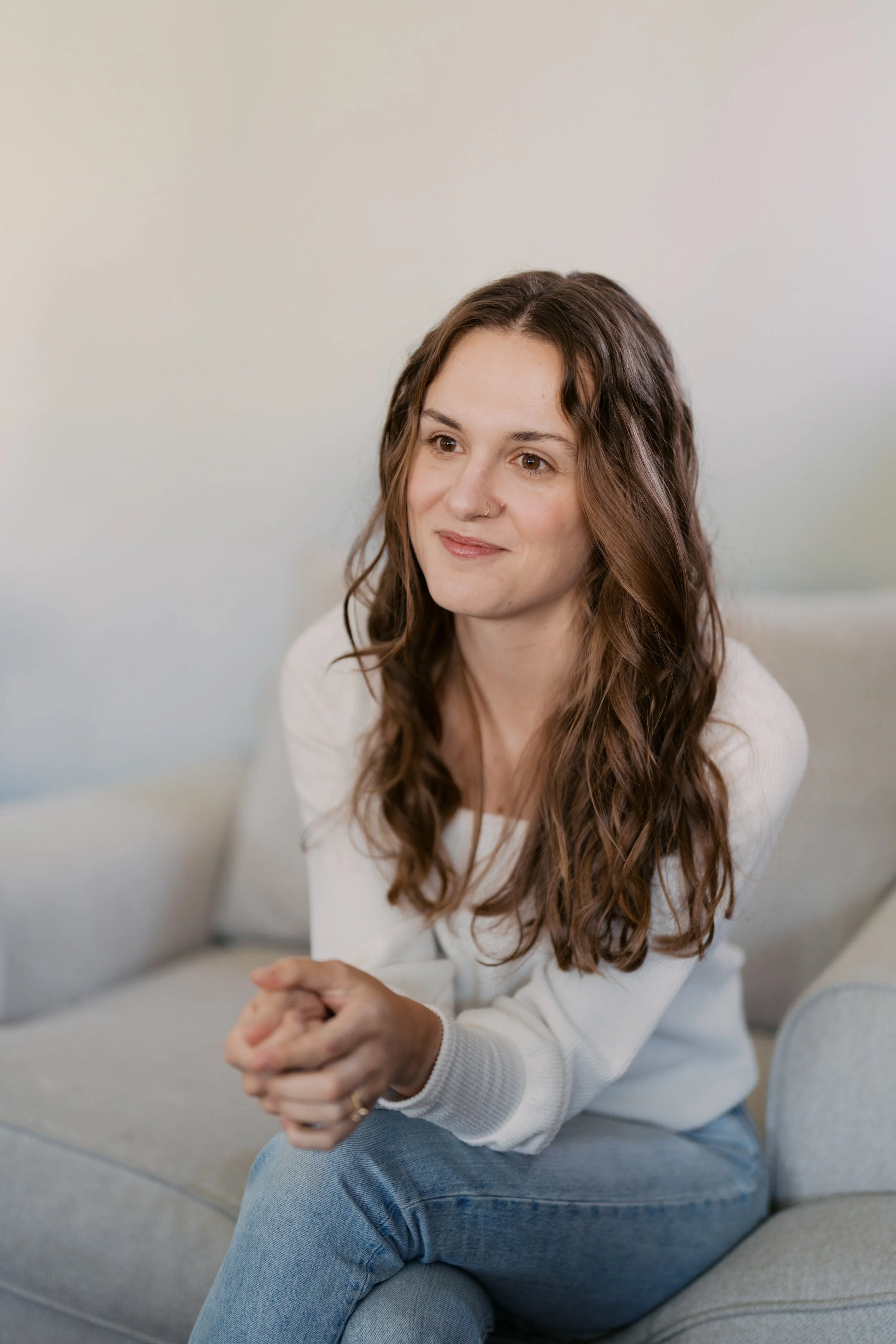 A young woman with long wavy brown hair sitting on a light-colored sofa with her hands clasped, wearing a white long-sleeve shirt and jeans, smiling softly at the camera.