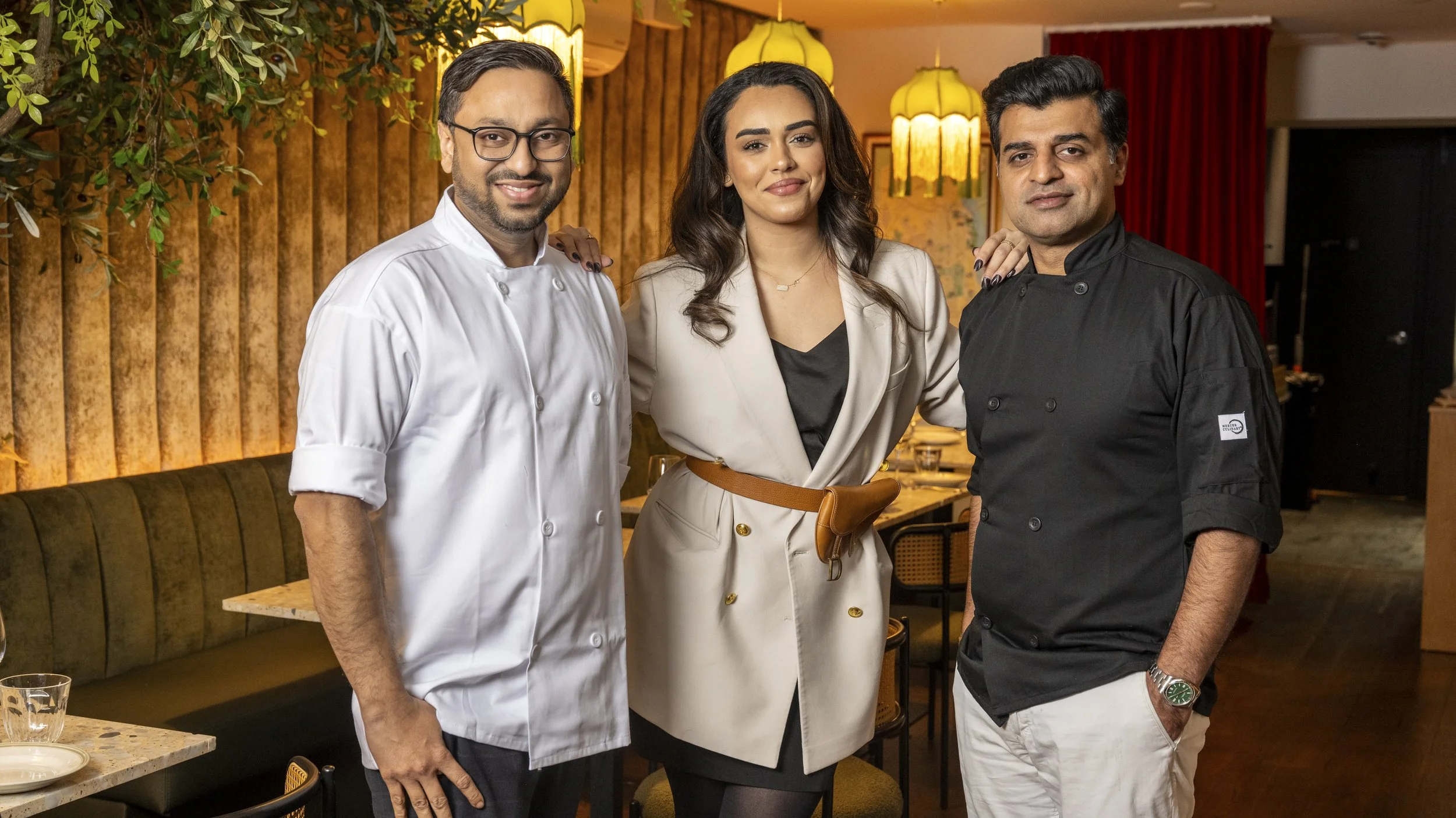 Three people, two men and one woman, standing together in a restaurant. They are dressed in professional chef and restaurant attire, with warm lighting and a cozy dining area in the background.
