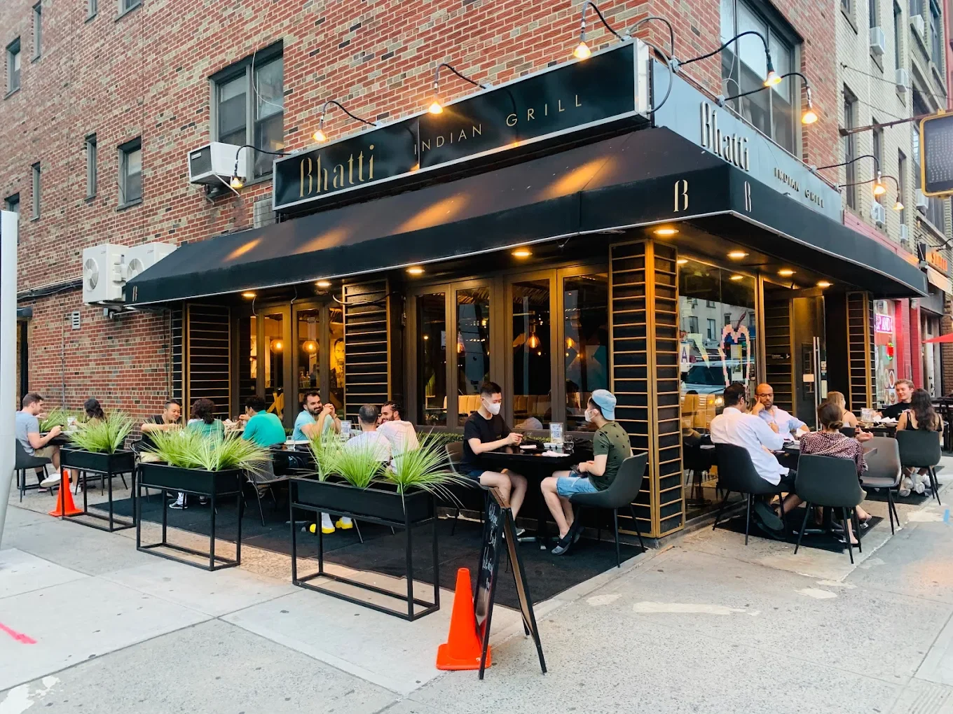Outdoor dining scene at Bhatti Indian Grill with people sitting at tables outside on the sidewalk, some wearing masks, in front of a brick building with a black awning and sign.