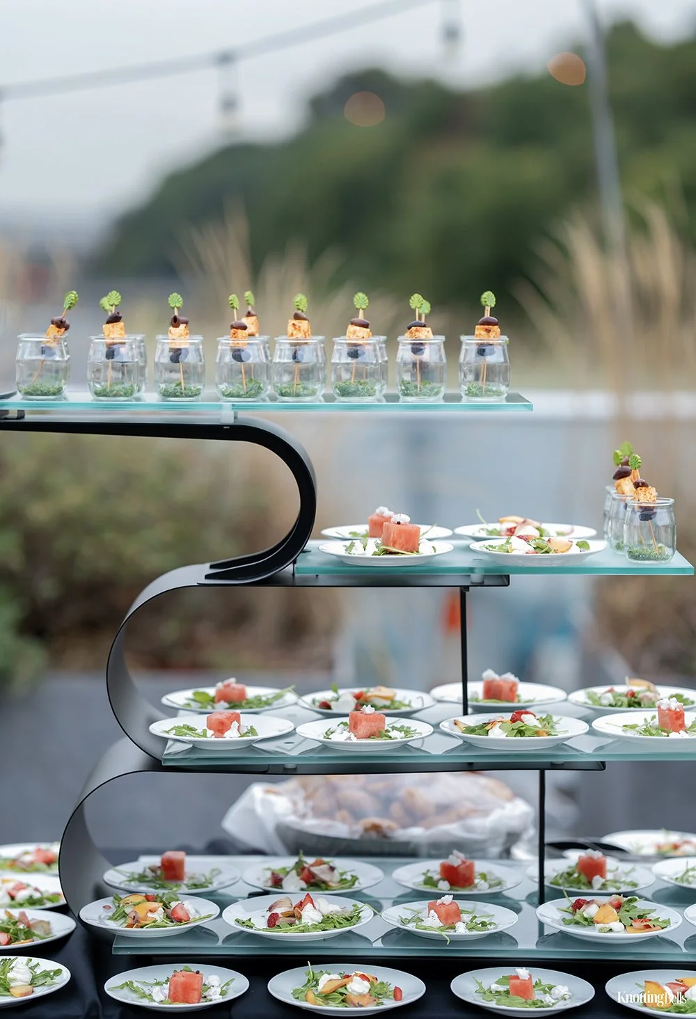 Elegant multi-tiered food display with small appetizer plates, salads, and garnishes, set outdoors with blurred natural background.