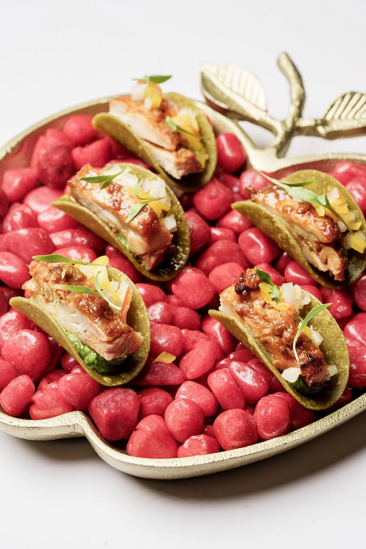 Five green chili peppers stuffed with chicken, cheese, and salsa, garnished with chopped onions and herbs, on a bed of pink radish balls in a decorative golden dish.