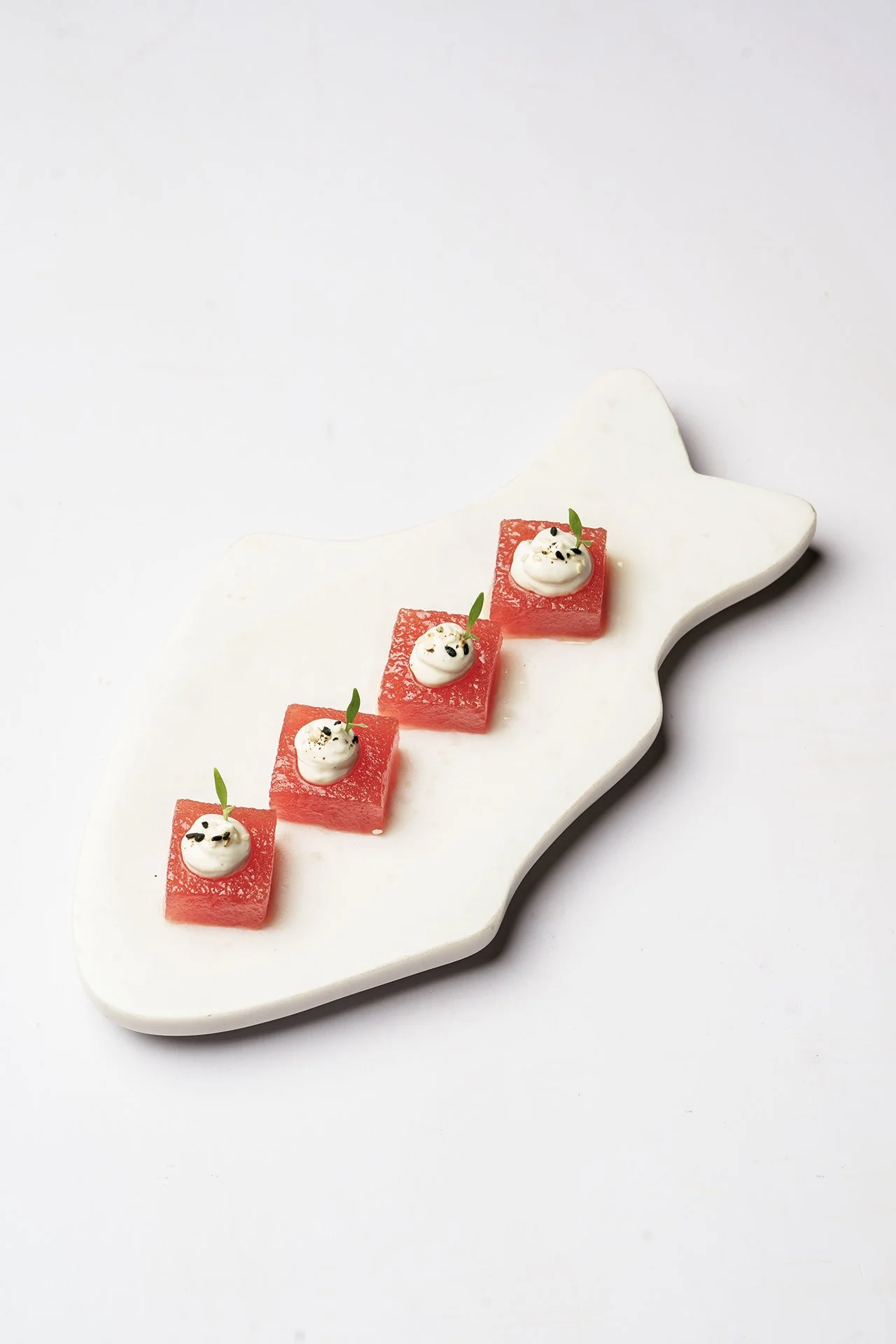 Four pieces of watermelon topped with cream and garnished with black sesame seeds and small green leaves, arranged on a white fish-shaped plate.