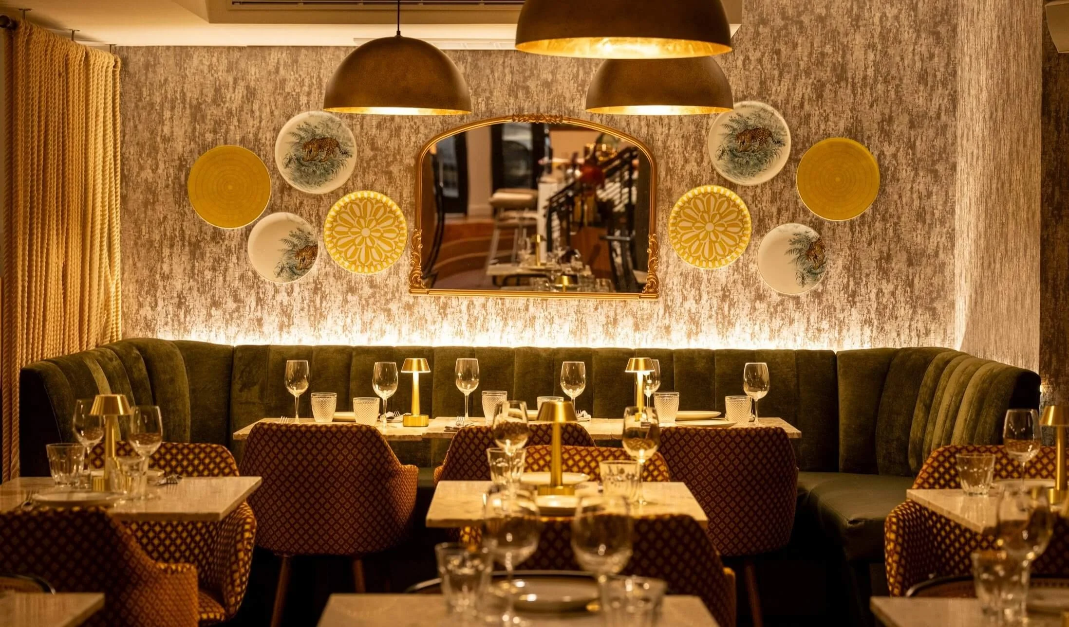 Elegant restaurant with a wall decorated with plates and a mirror, a green velvet banquette, and tables set with wine glasses, water glasses, and gold lamps. Dilli Dilli NYC.